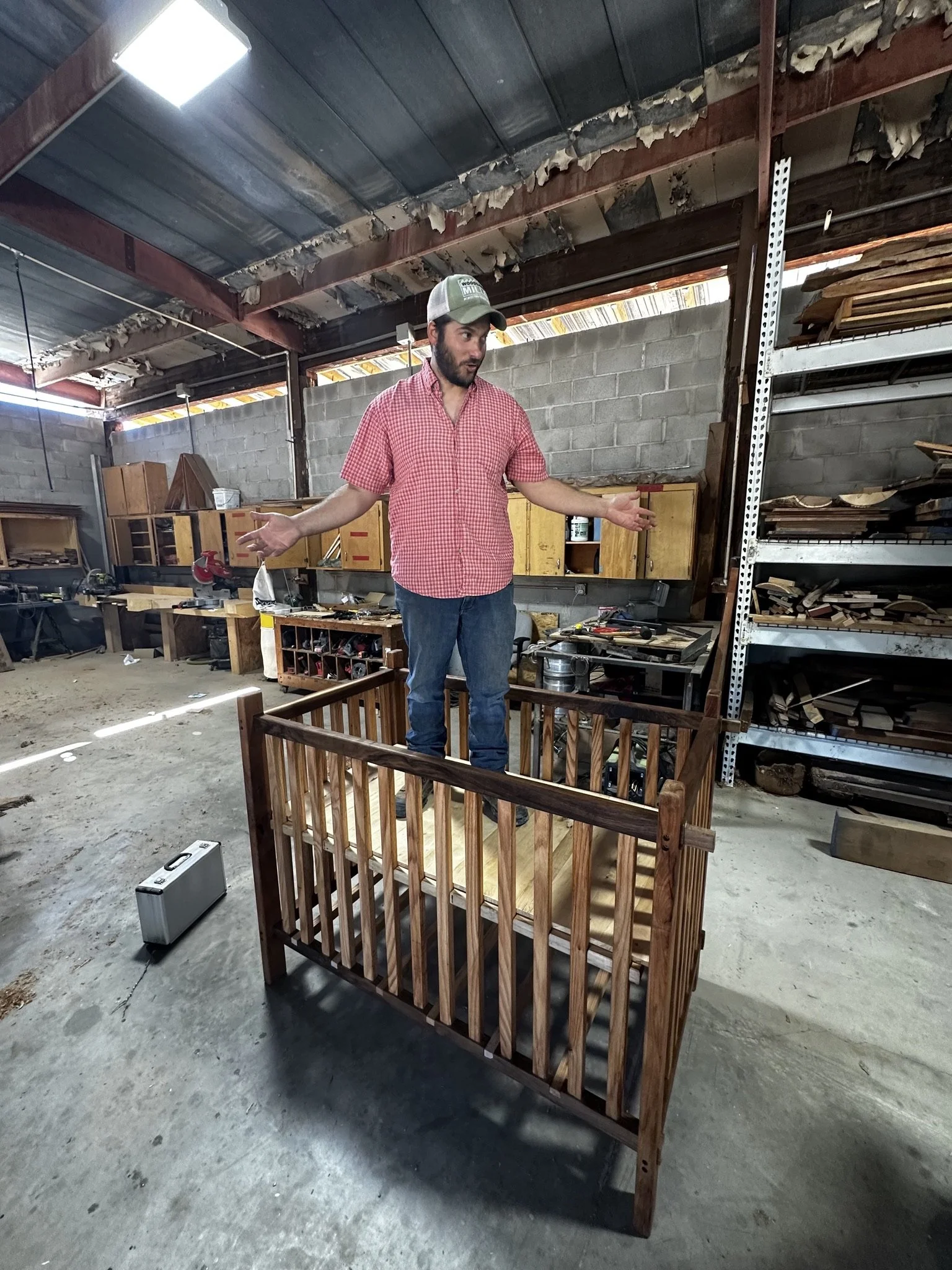 A man standing inside a moving crib made of wood in a woodworking workshop, gesturing with his arms out.