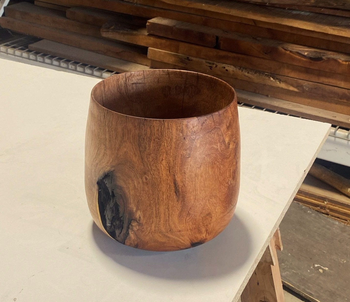 A wooden bowl with a smooth, rounded shape and natural grain patterns, placed on a white surface in a woodworking workshop.