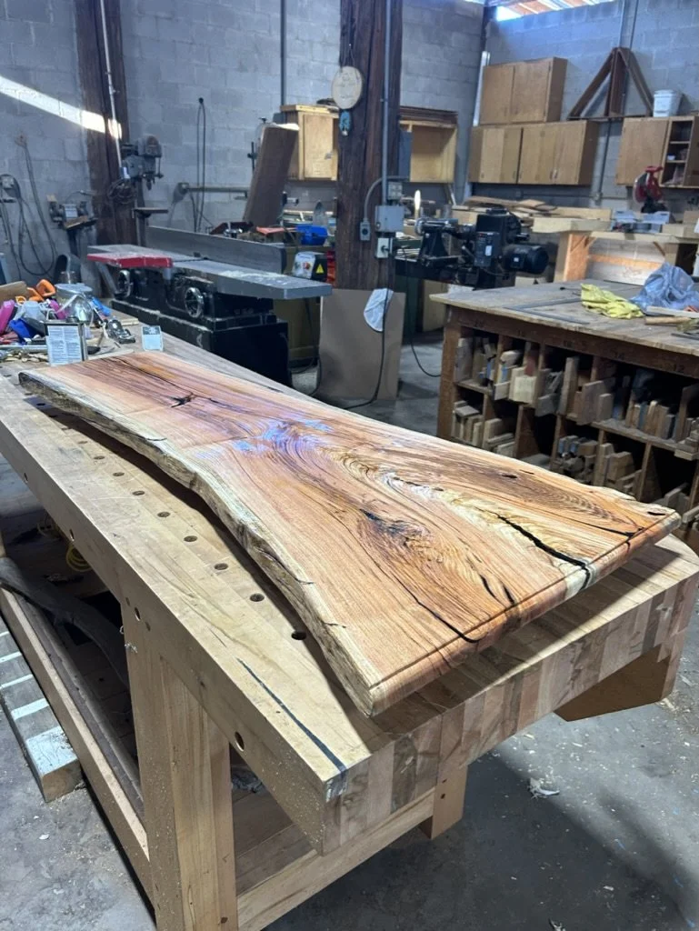 A large, polished piece of natural wood with visible grain and knots, resting on a sturdy woodworking workbench inside a workshop with woodworking tools, equipment, and storage cabinets in the background.