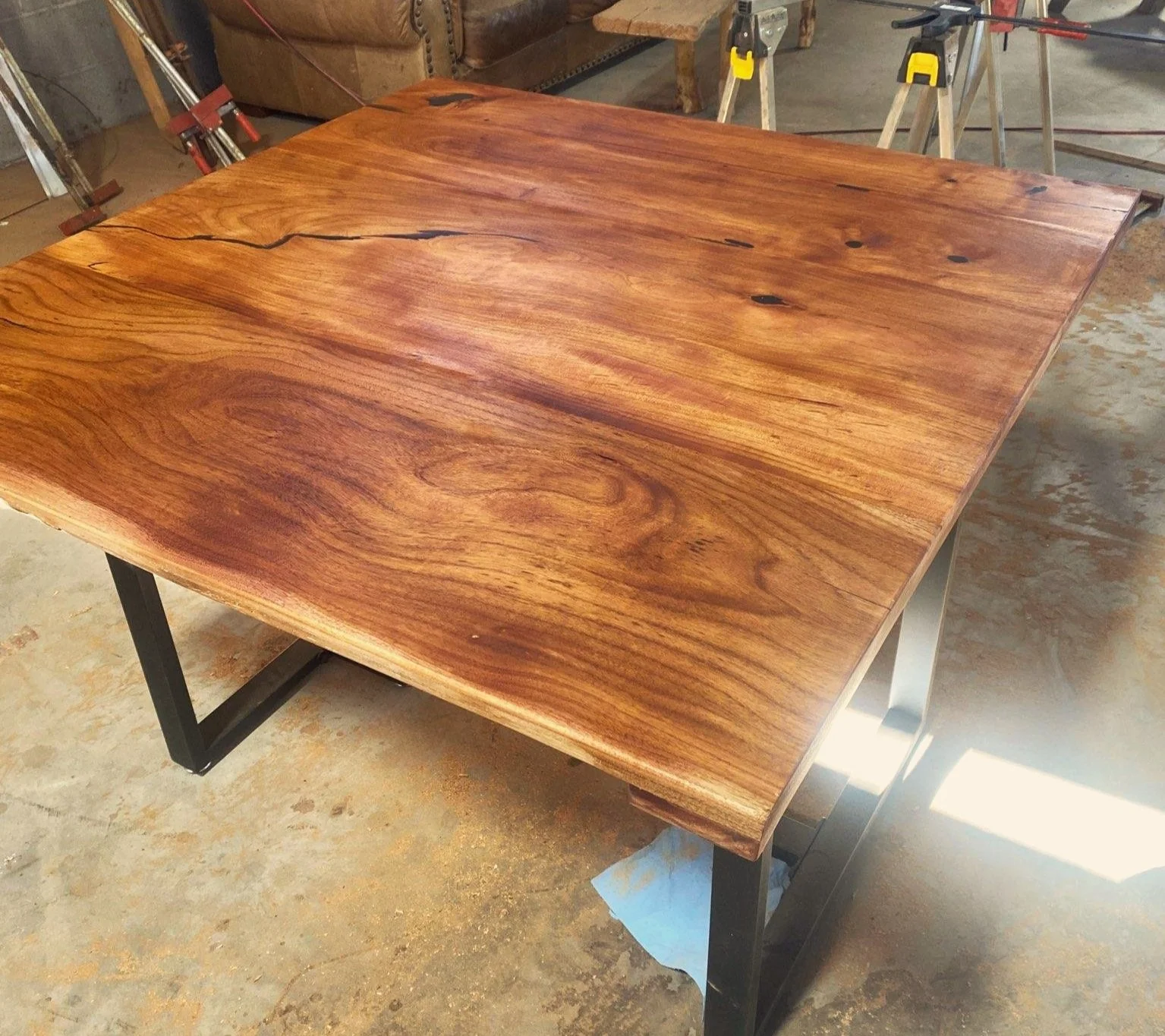 A wooden table with a natural finish showing wood grain and knots, supported by black metal legs, in a workshop setting.