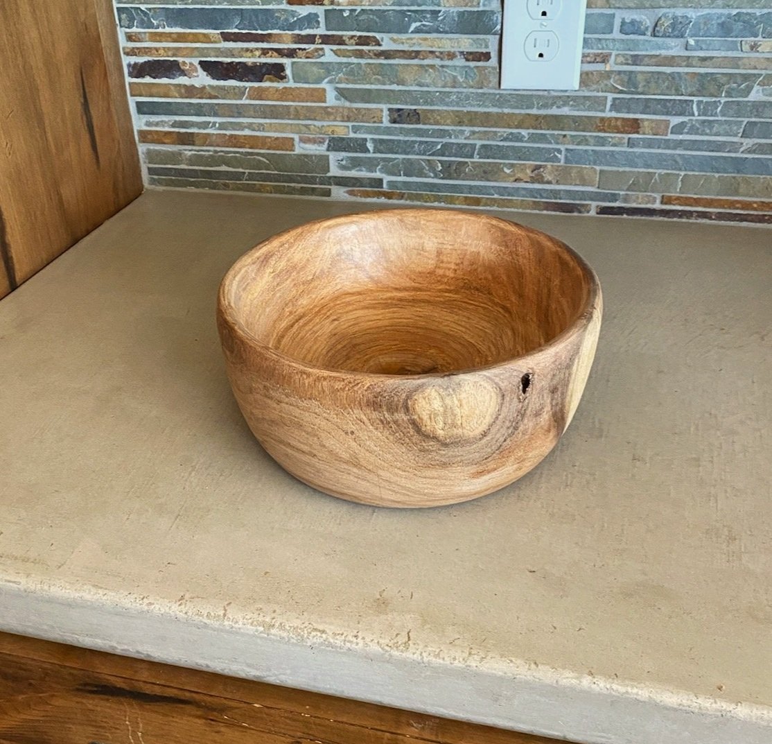 A round wooden bowl on a beige countertop against a brick and wood wall with electrical outlets.
