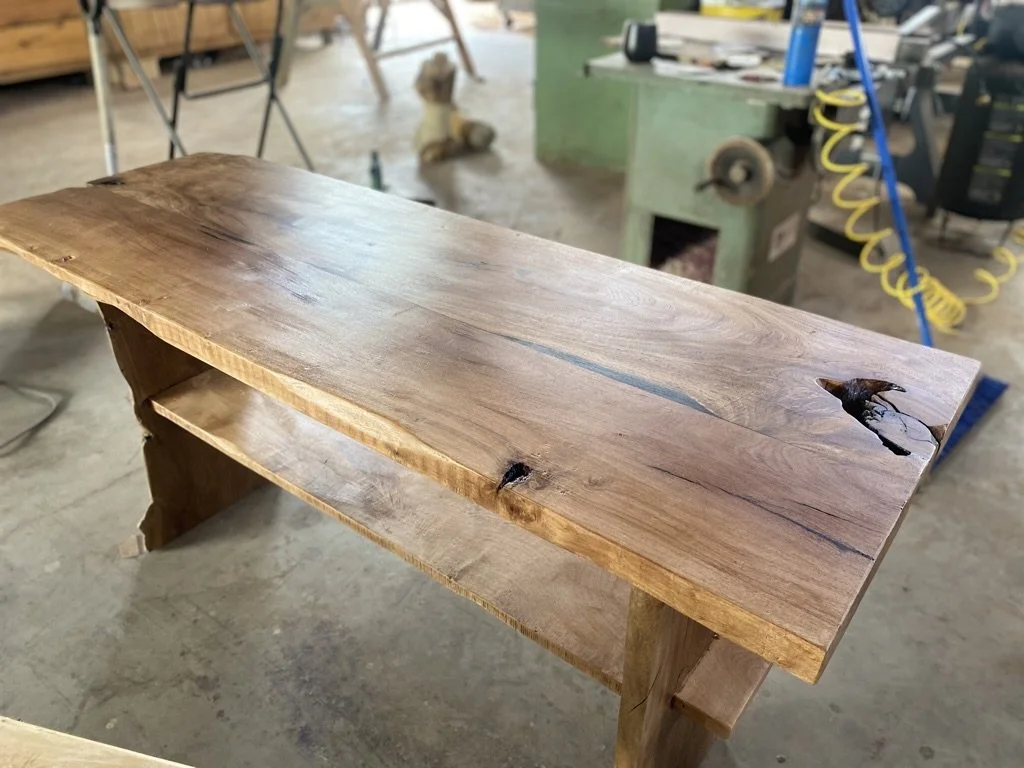 A wooden table in a woodworking shop, with a live edge and a knot hole, surrounded by woodworking tools and equipment.