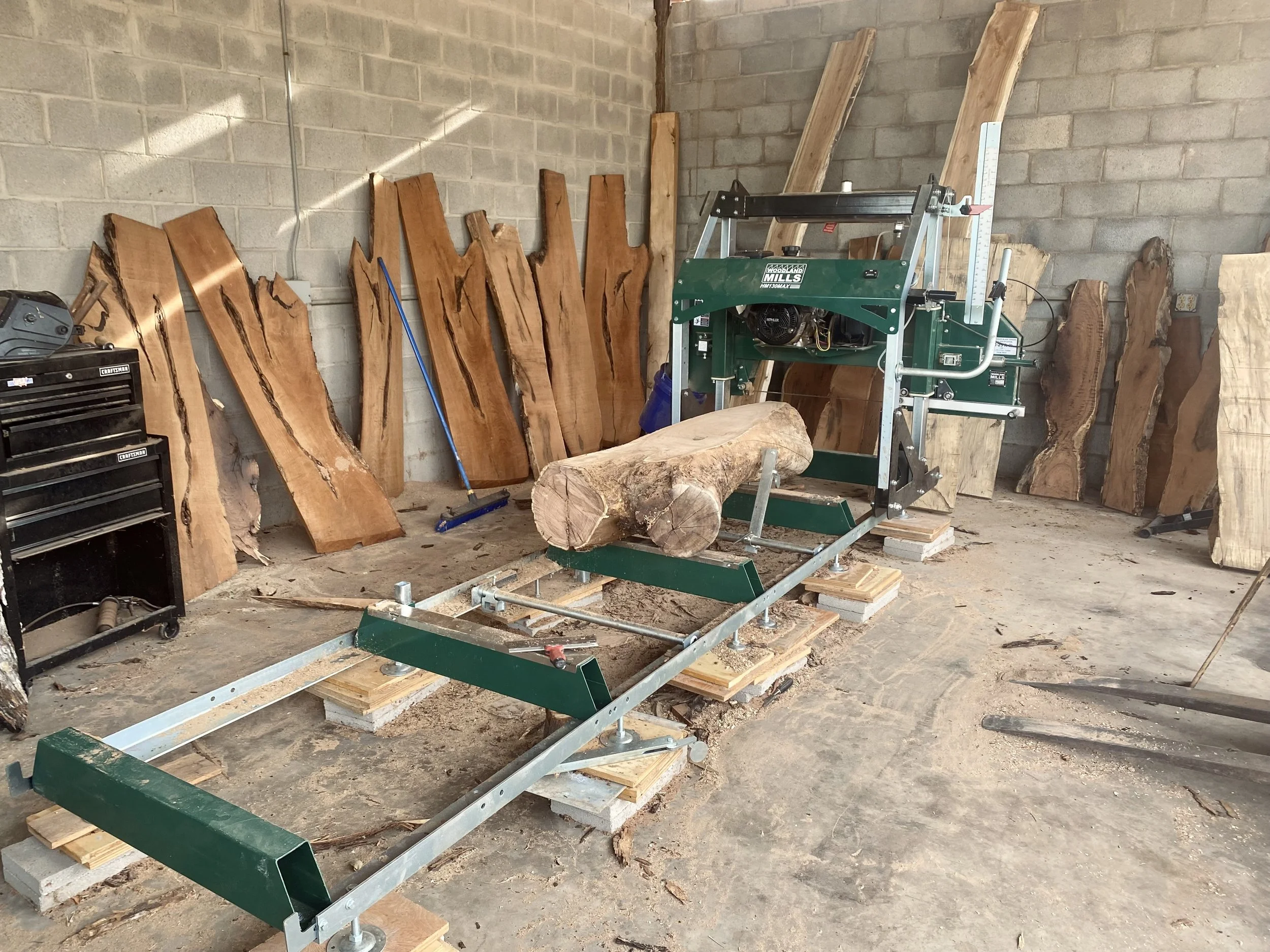 Woodworking shop with a large bandsaw cutting a log into slabs, surrounded by various slabs of wood against a brick wall.