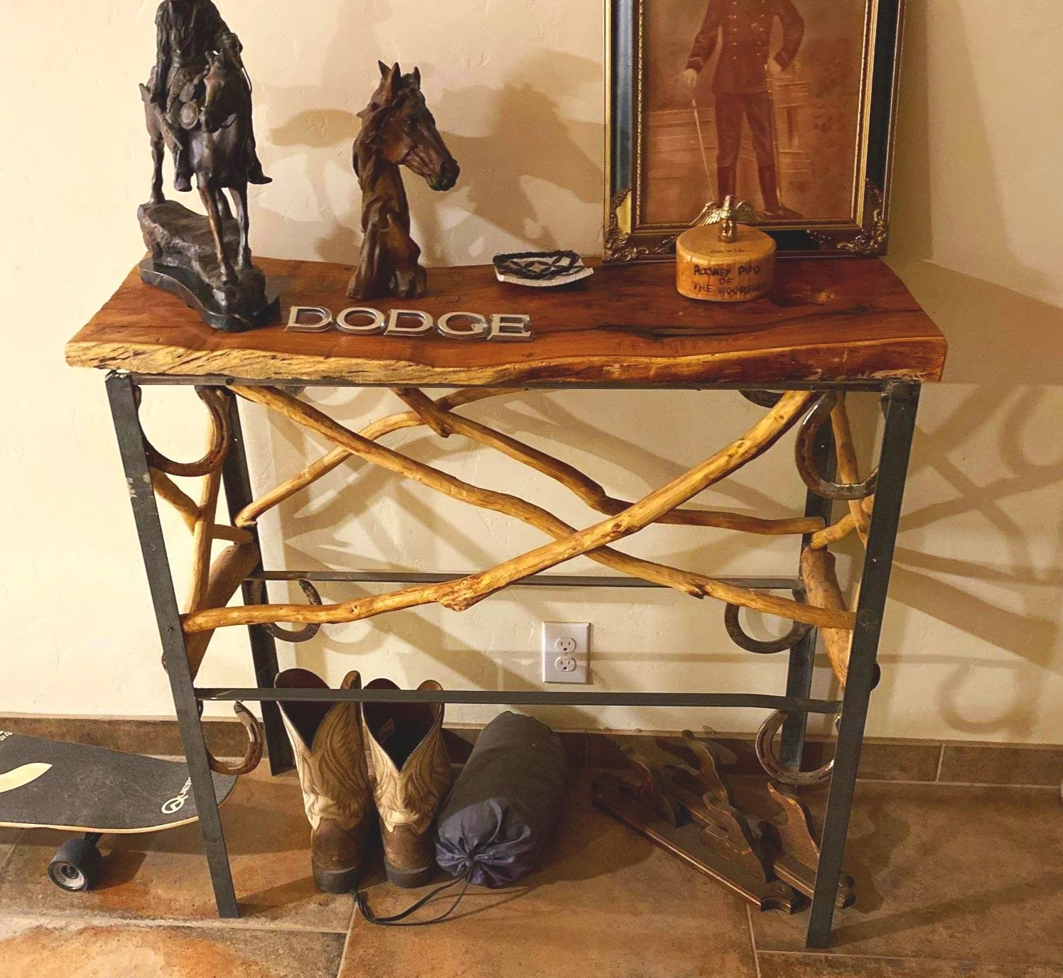 A rustic wooden and metal console table with horse figurines, a framed portrait, and various decor items on top; cowboy boots and a rolled sleeping bag on the floor nearby; skateboard leaning against the wall.