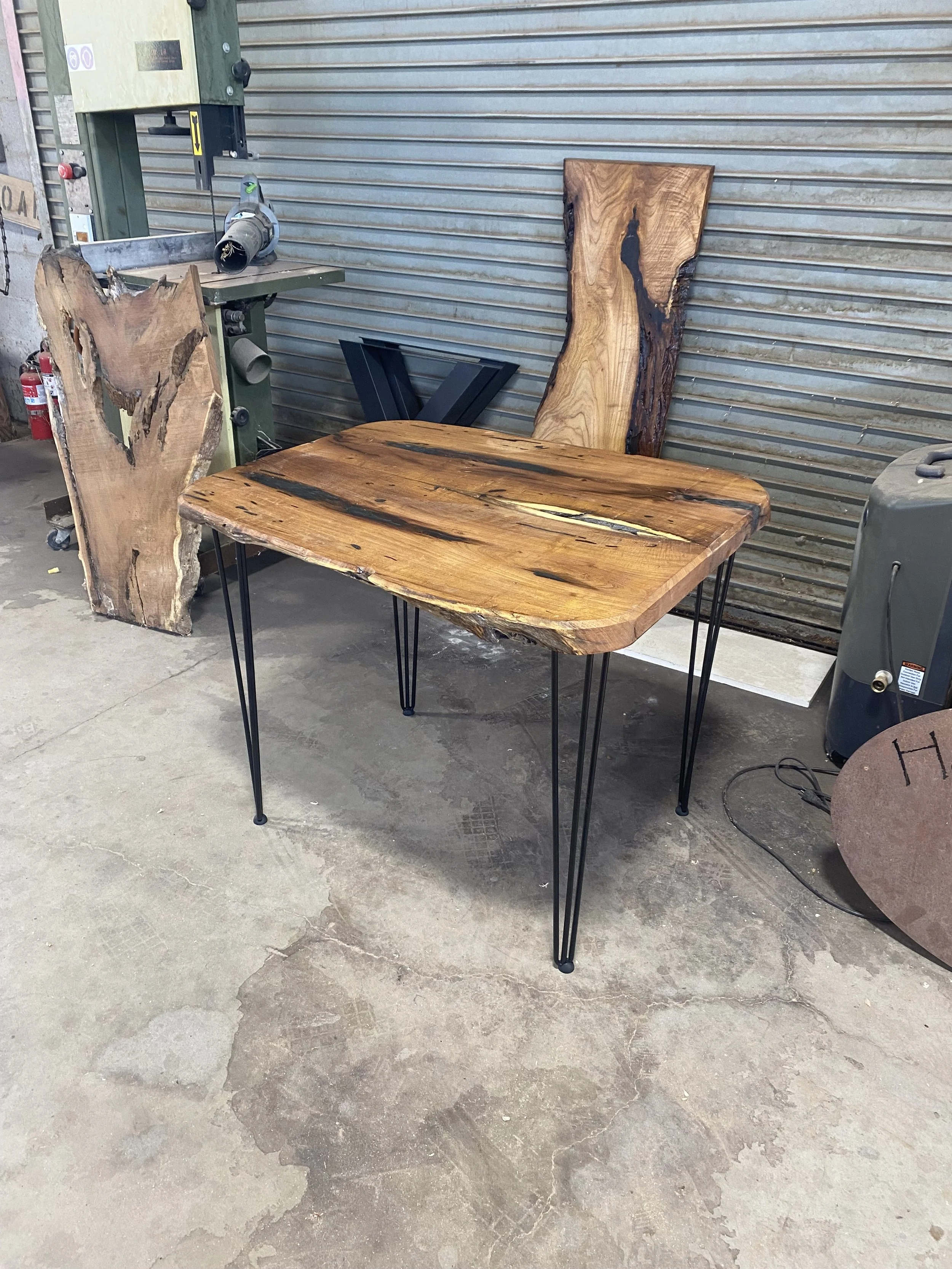 A woodworking workshop with a large, polished wooden table supported by black metal hairpin legs, surrounded by three large wooden slabs, a band saw, a fire extinguisher on a concrete floor, and a metal roller shutter in the background.