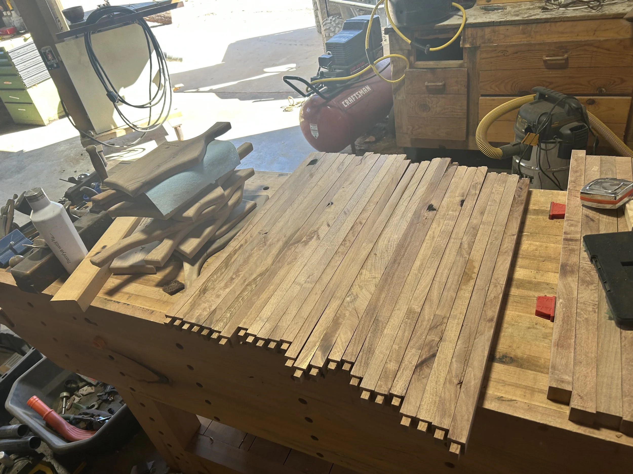 A workbench in a garage with a wooden top and various tools, including a cordless drill, a large orange air compressor, a small tool box, and some wooden pieces, with natural light coming from outside.