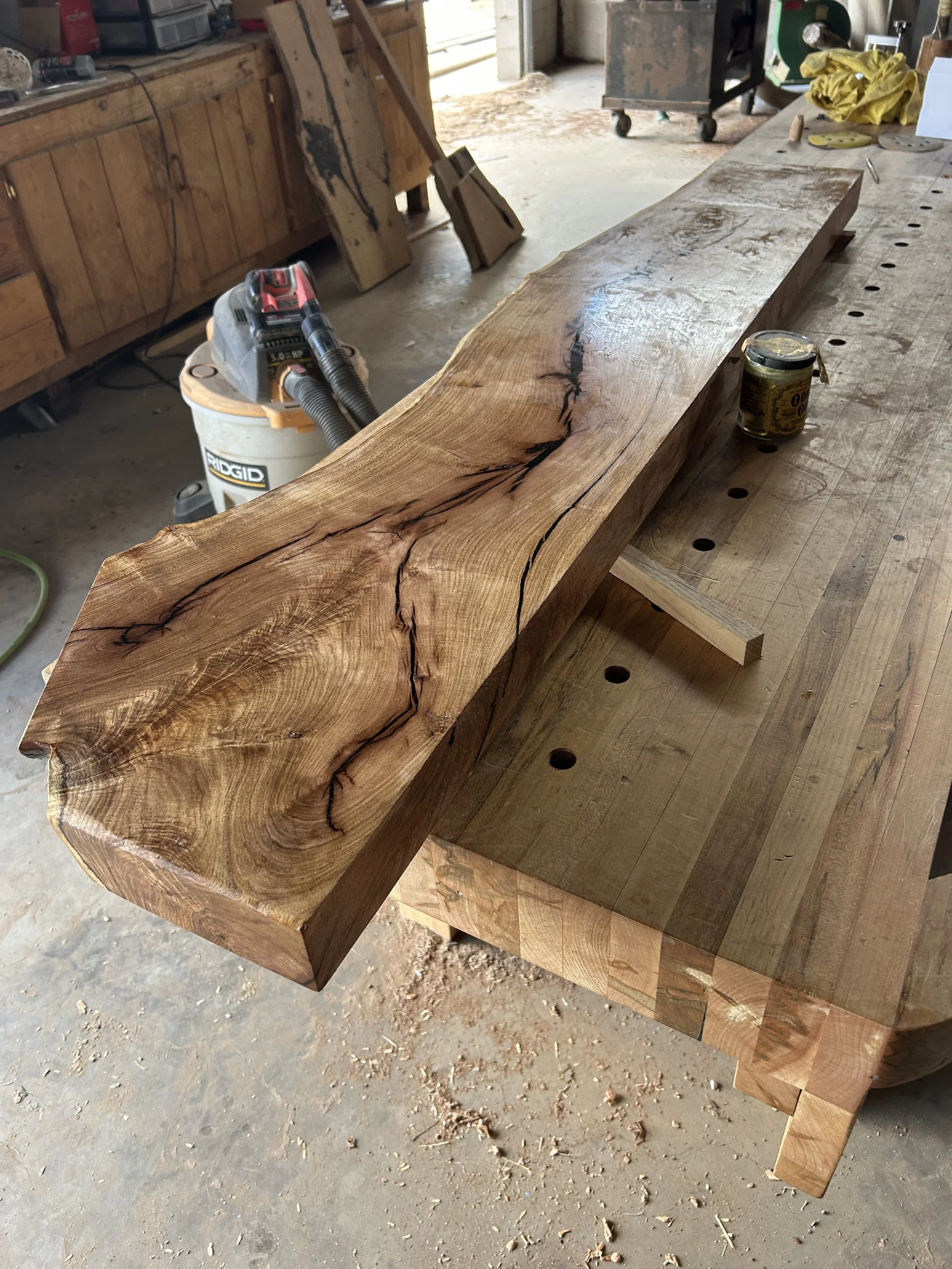 A rustic wooden table in a woodworking shop with various tools and materials on and around it, including a can of wood finish and a shop vacuum, with sawdust and wood shavings on the floor.