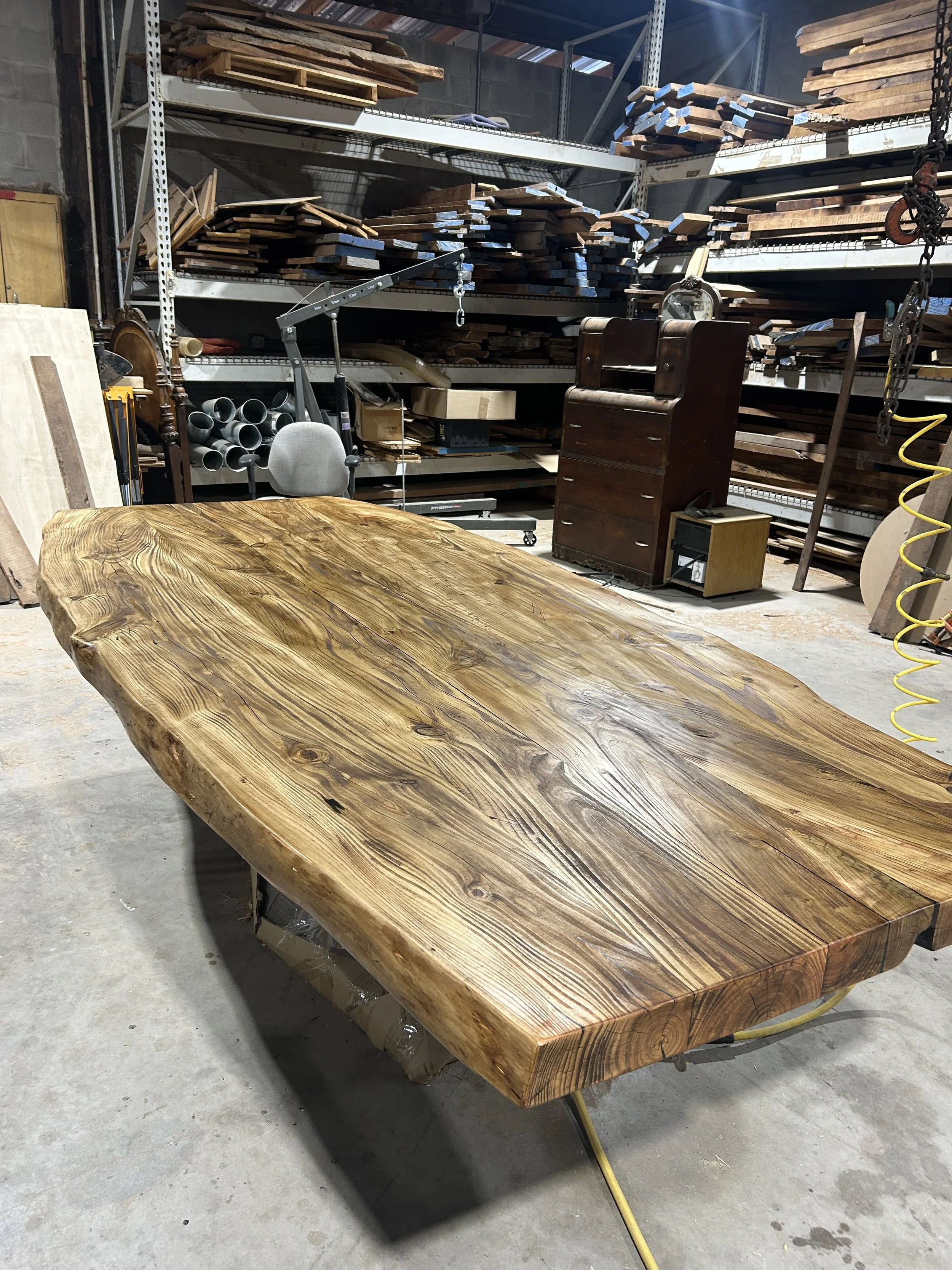 A large, polished wooden slab with a natural, uneven edge placed on a metal stand in a woodworking shop or storage area. In the background, shelves are filled with various lengths of cut wood and woodworking tools. The space appears industrial with a