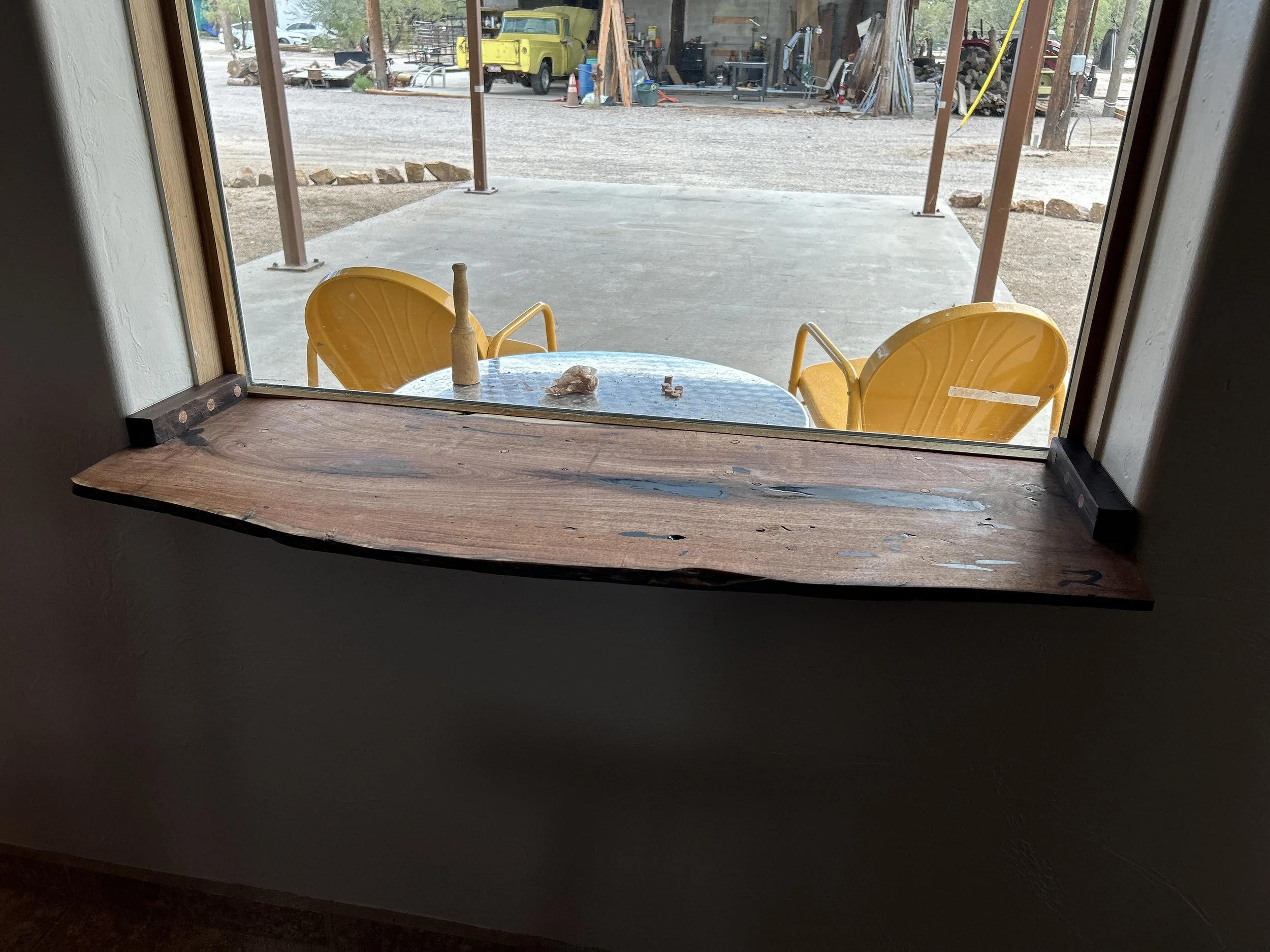 View through a window showing a covered patio area with yellow outdoor chairs and a small round table. Outside, there is a gravel lot with various items, including a yellow truck, trees, and a woodpile in the background.