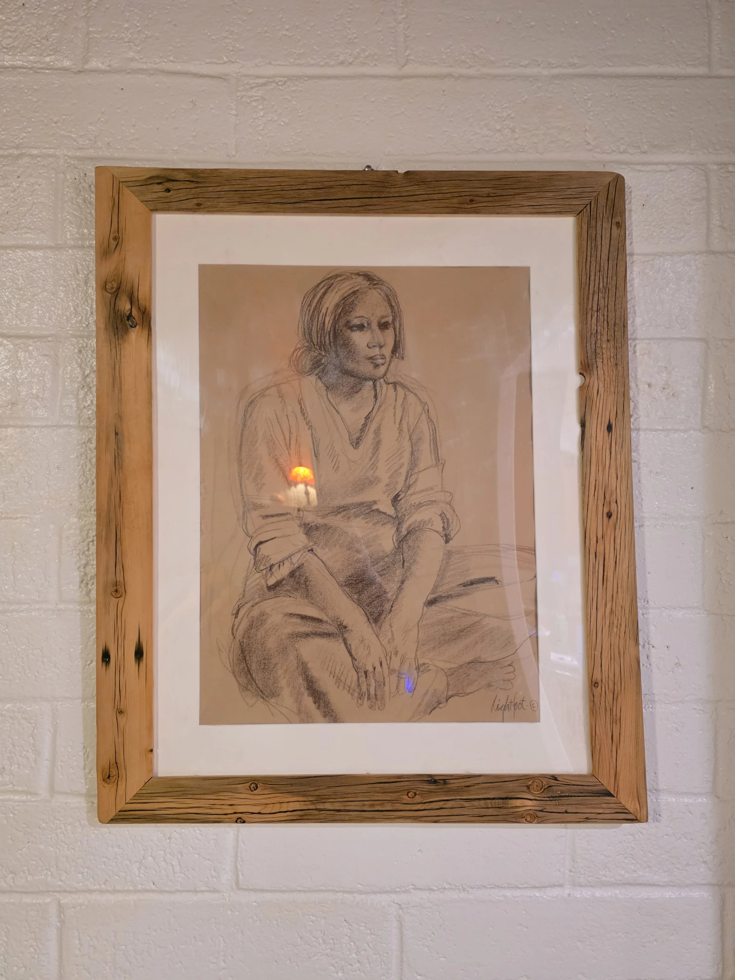A framed sketch of a seated woman with short hair, sitting with her legs crossed, on a textured white wall.