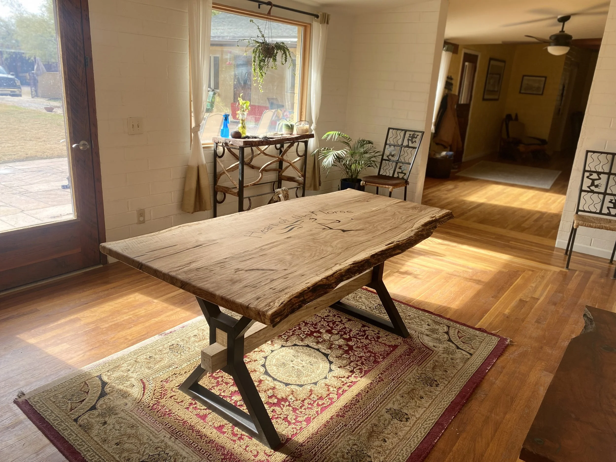 A wooden table with metal legs sits on a patterned area rug in a room with hardwood flooring. Light streams in through a large window with cream curtains, with plants and decorative items on a table beneath the window. There are chairs with metal fra