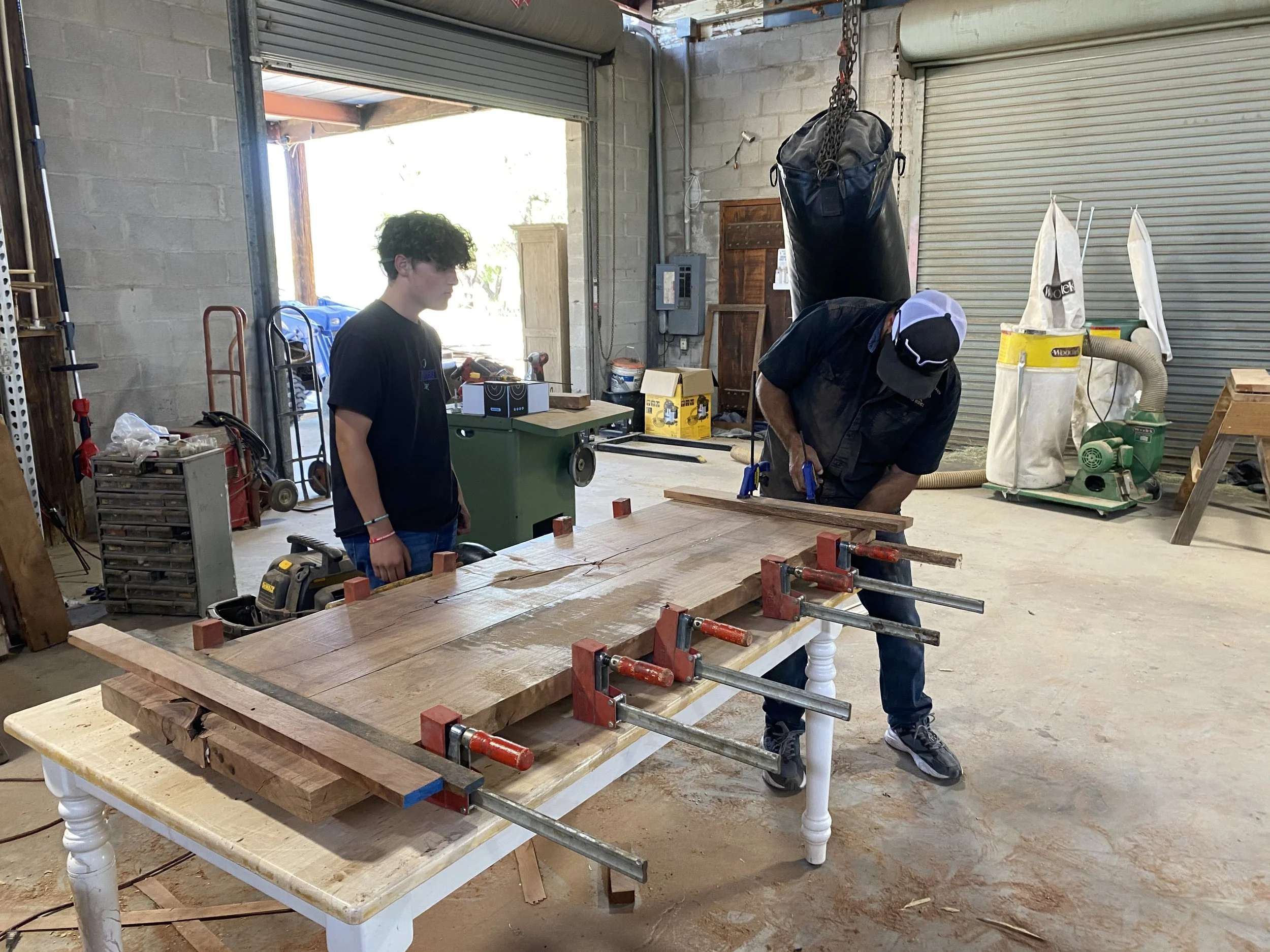 Two people working in a woodworking shop, one man is bending over a table, the other standing nearby, with woodworking tools and equipment around.