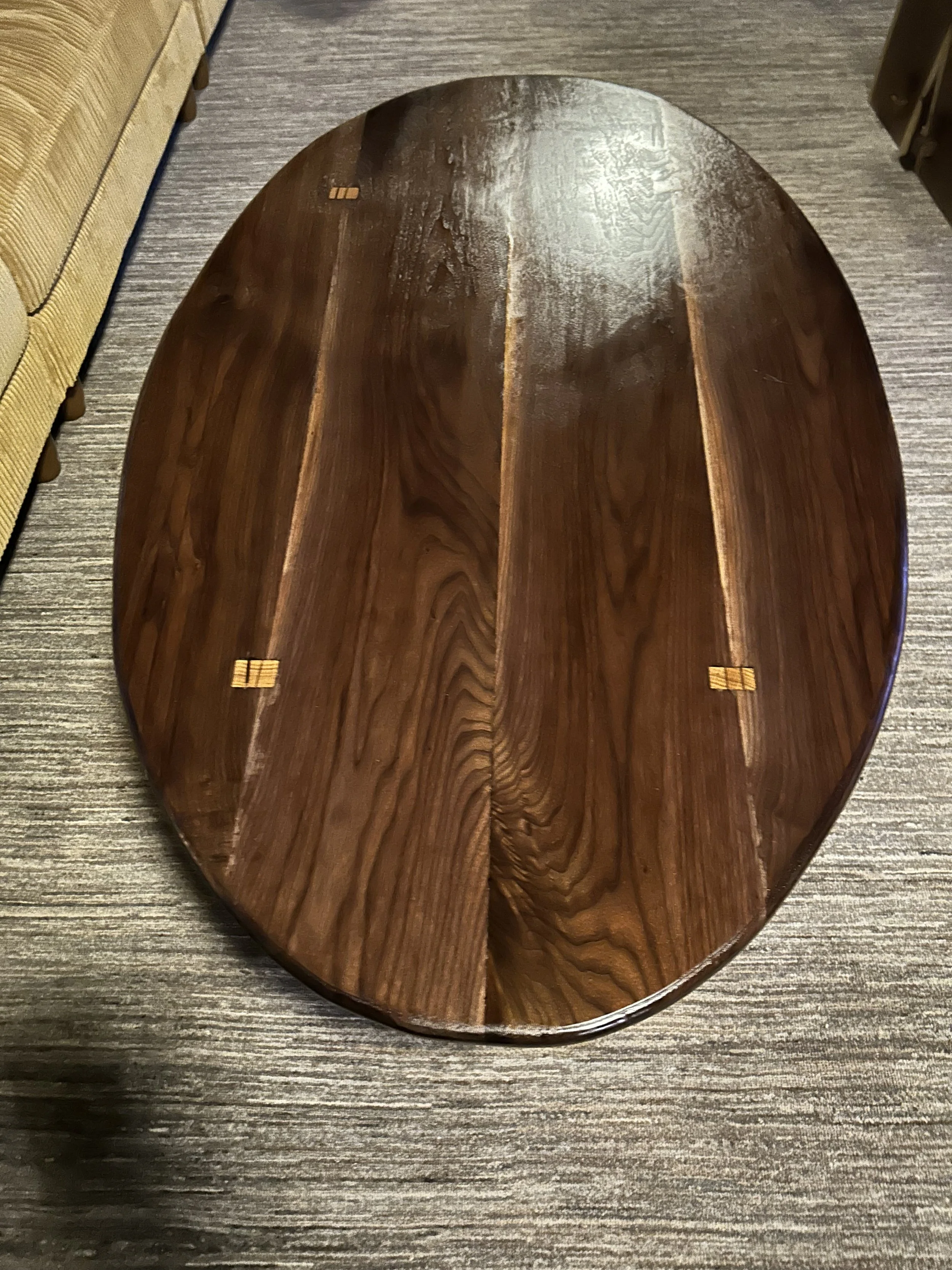 A wooden oval table with a glossy finish, positioned on a gray textured floor, with part of a yellow upholstered piece of furniture visible on the left.