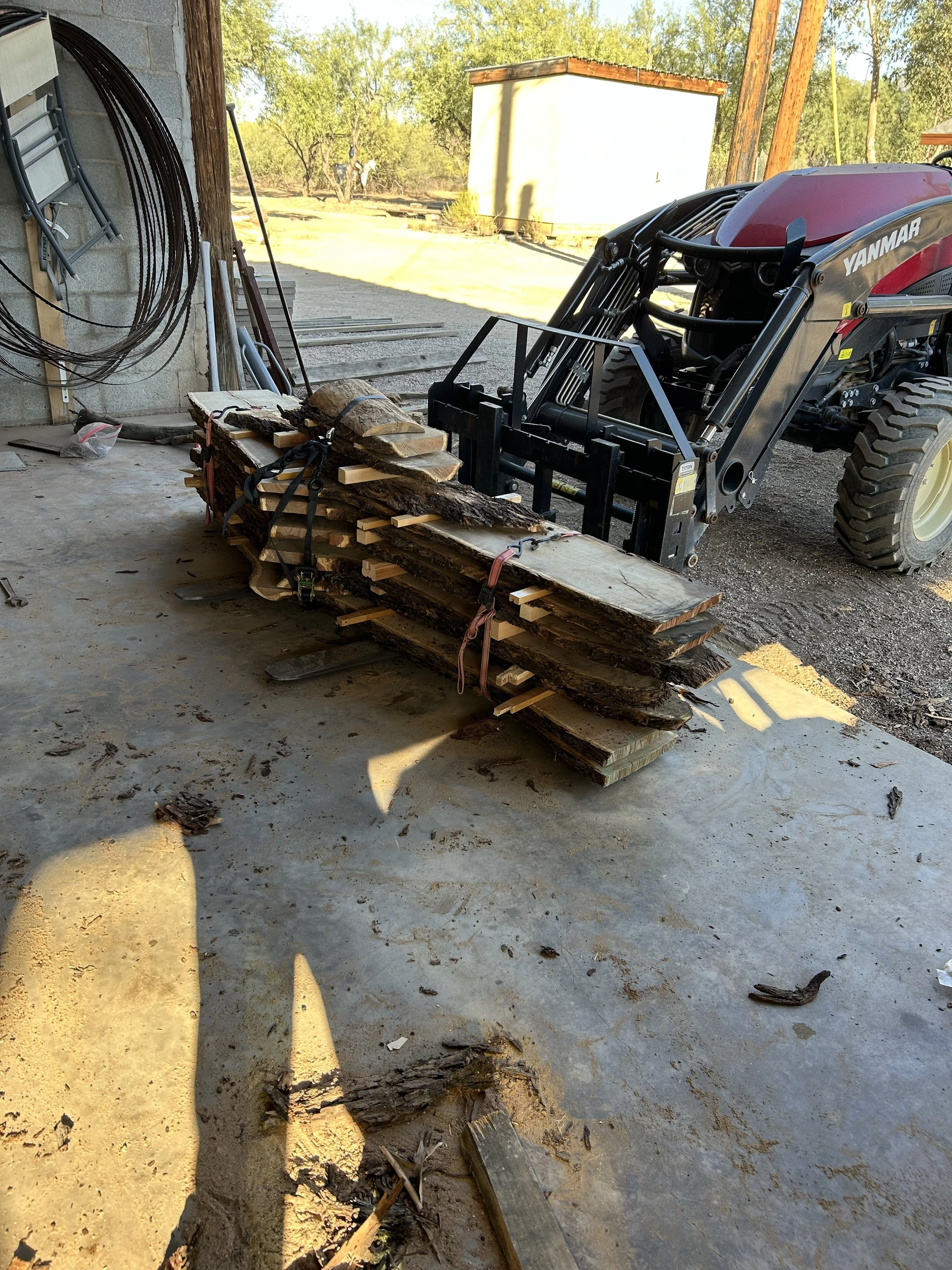 A stack of cut firewood on a wooden platform next to a black tractor with a front loader attachment indoors beside a concrete wall, with outdoor trees and a small building visible in the background.