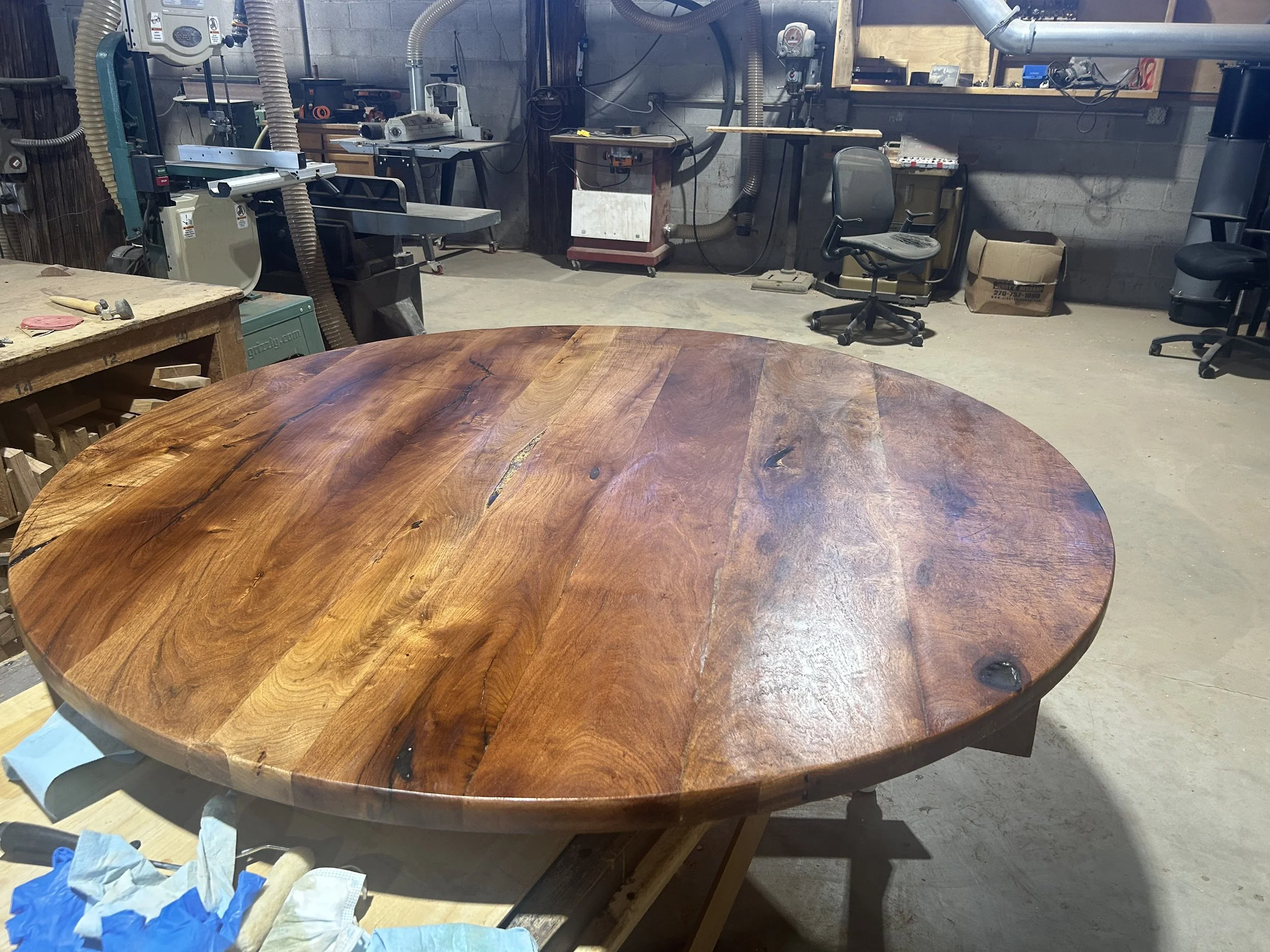 A large round wooden table with a polished surface in a workshop. The background shows woodworking tools, equipment, and chairs, indicating an active woodworking or furniture-making space.