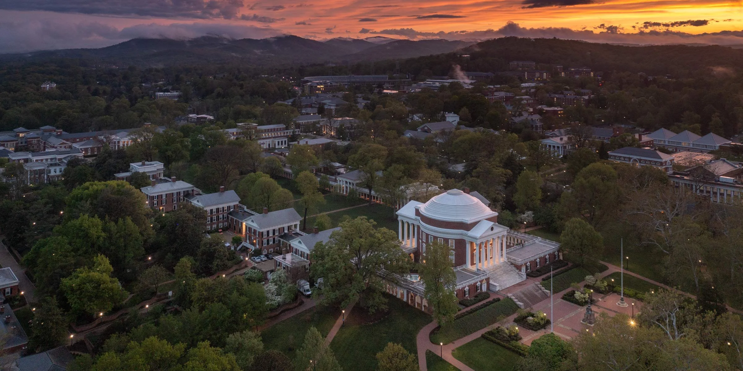 An aerial view of a university campus at sunset with a prominent red brick building featuring white columns in the foreground, surrounded by lush green trees and other campus buildings, with mountains and colorful sky in the background.
