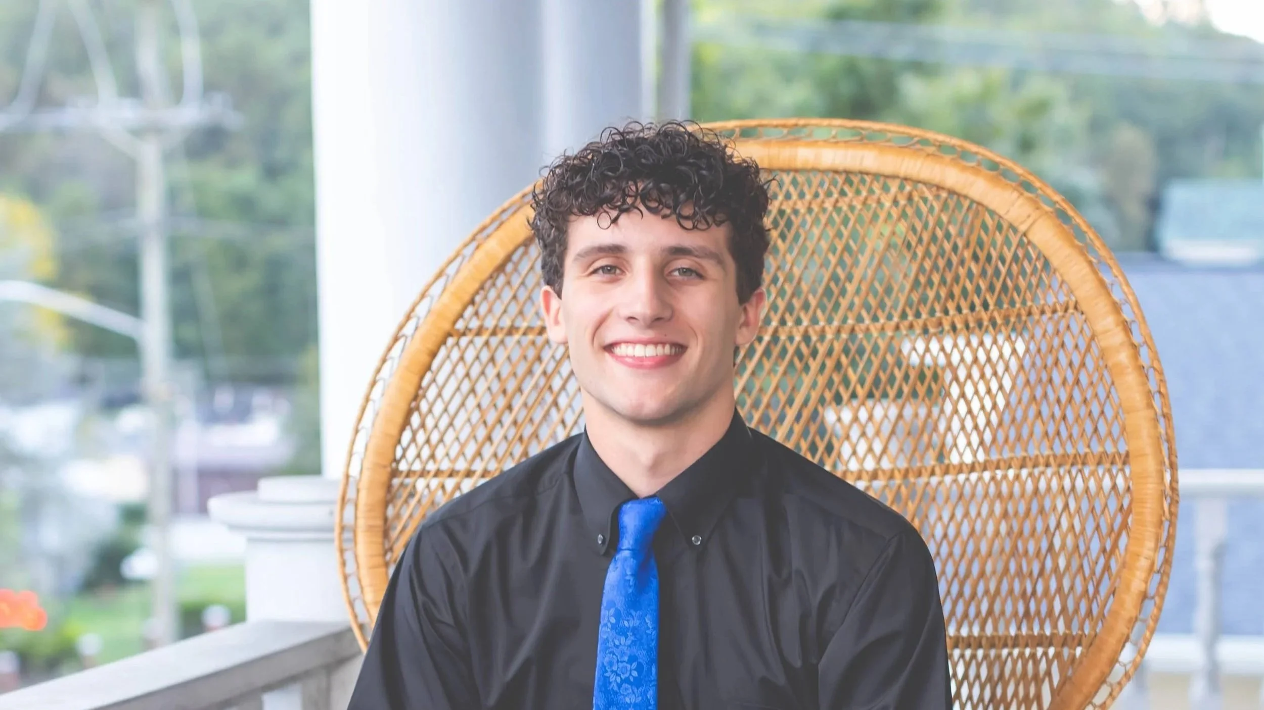 A young man with curly dark hair smiling, wearing a black shirt and blue tie, seated in a rattan chair in a bright indoor space with large windows and greenery outside.
