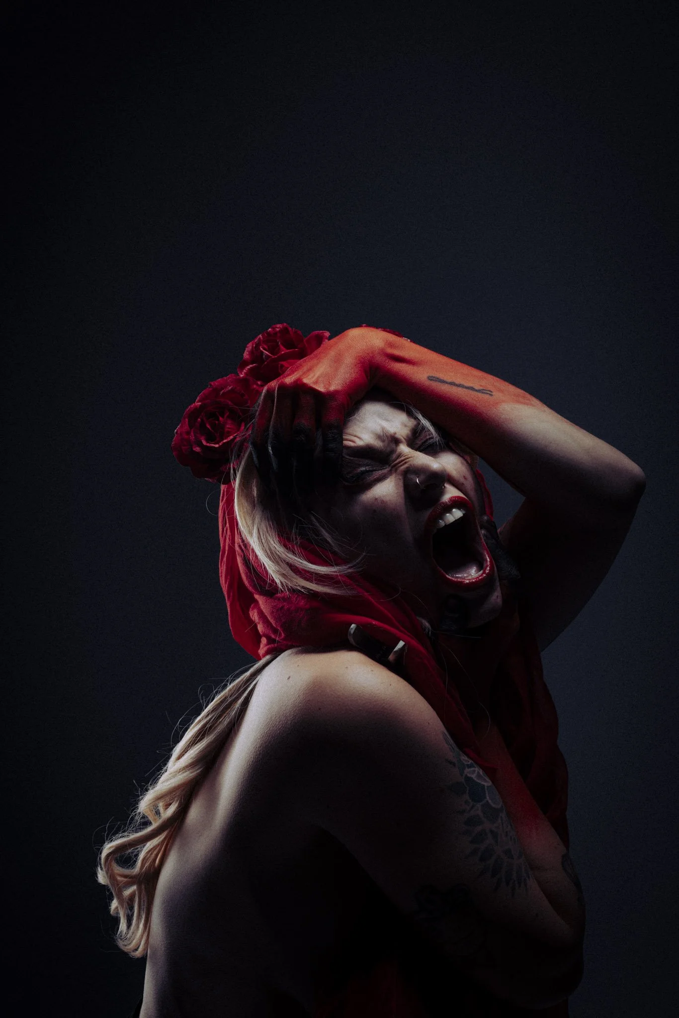 A woman with long hair, tattoo on her arm, wearing red gloves and a red headscarf, screaming or yelling, with her hand on her head, against a dark background.