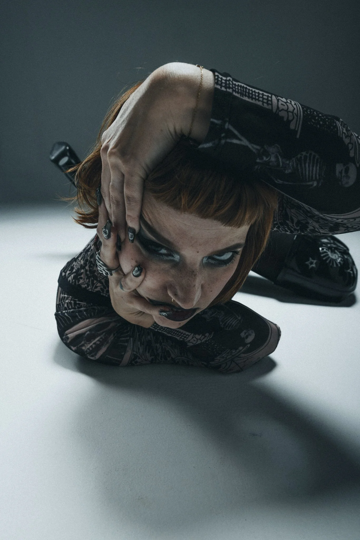 A person with short red hair lying on the floor, covering their face with one hand, wearing dark clothing with skeleton and skull patterns, and jewelry including rings and a necklace, against a plain white and gray background.