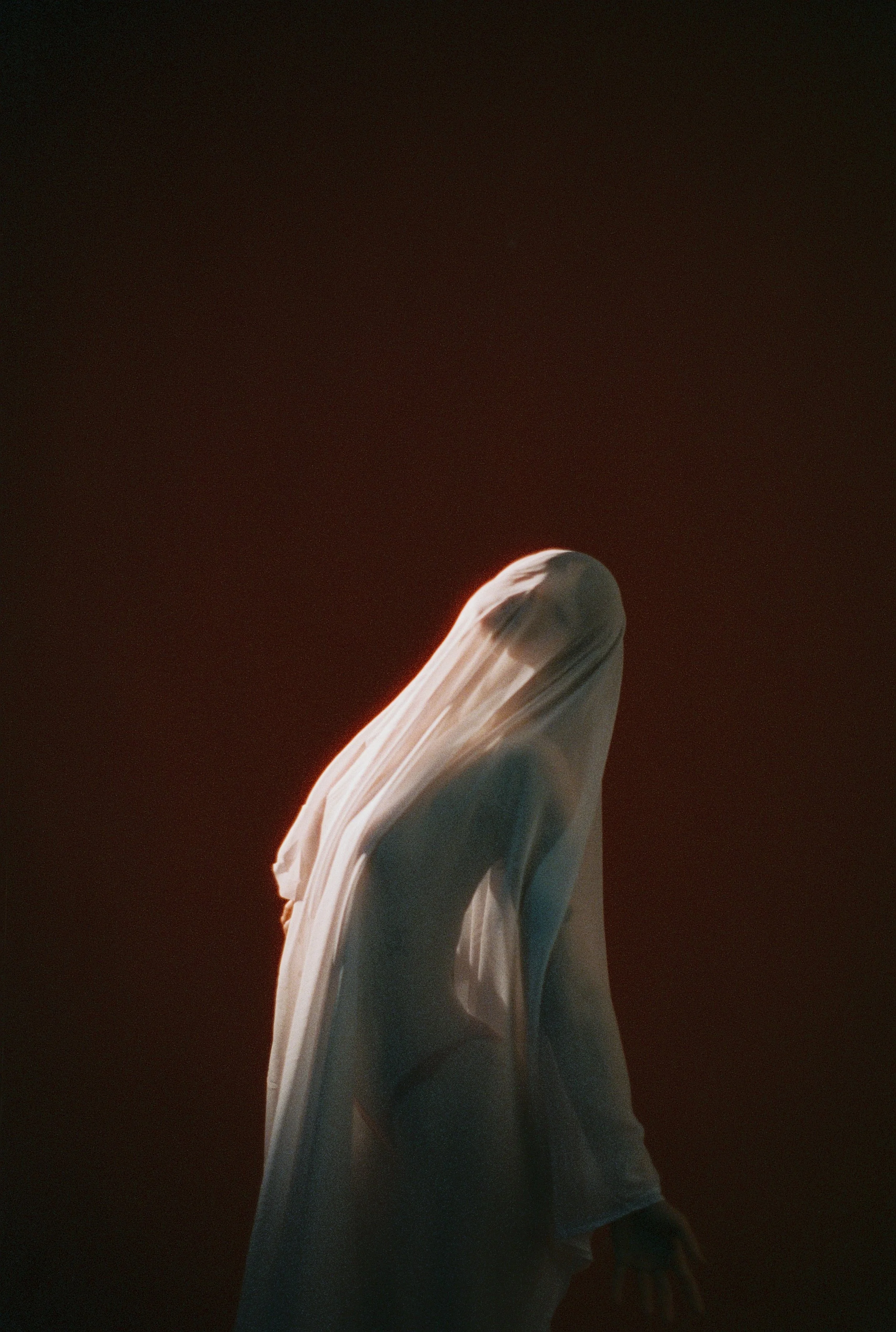 A person stands with their back turned, wearing a draped, flowing white garment. The background is dark, with a faint, warm light outlining the person's head and shoulders, creating a halo effect.