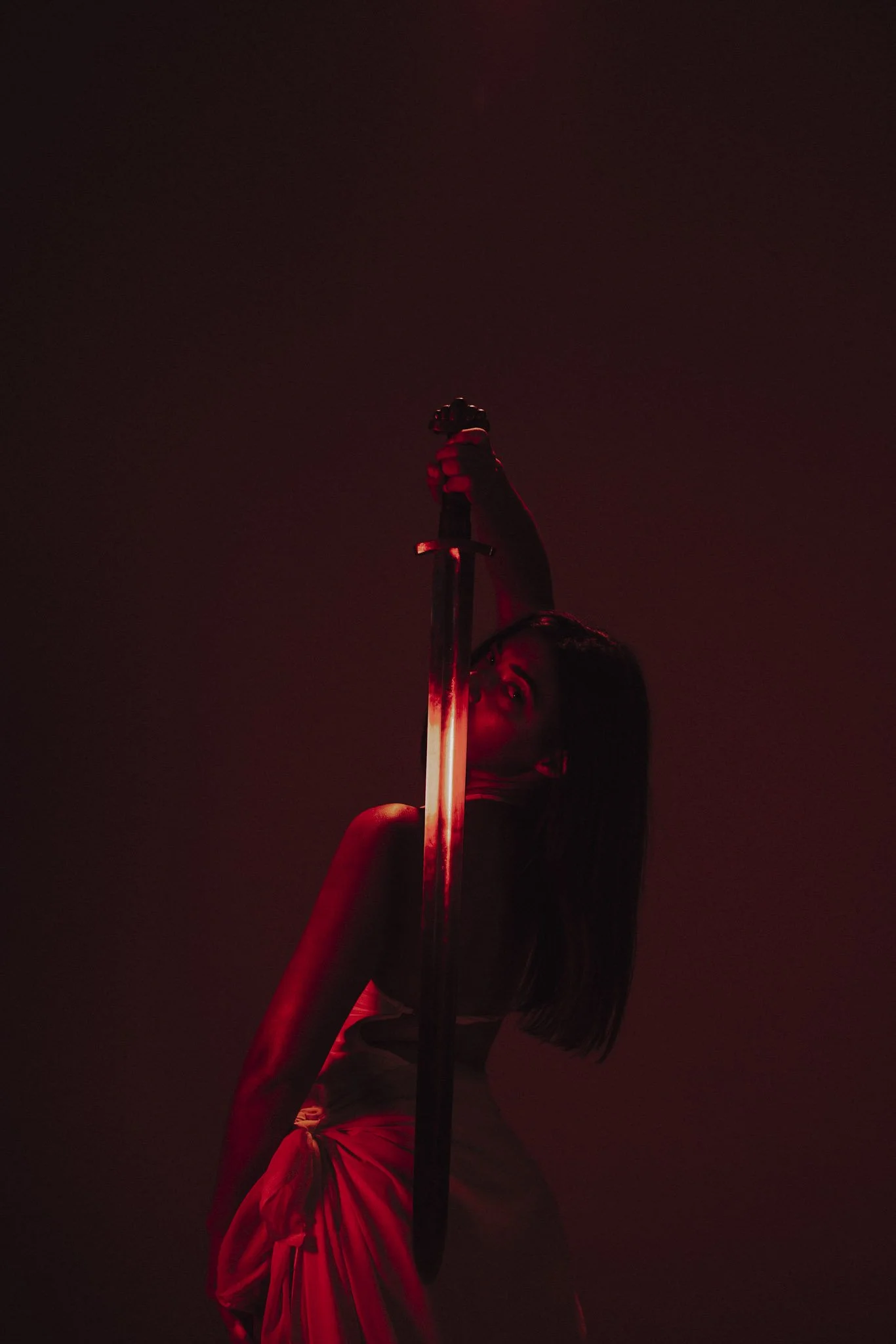 A person with dark hair and glasses is holding a sword vertically in the air, with a dark background illuminated by red light.