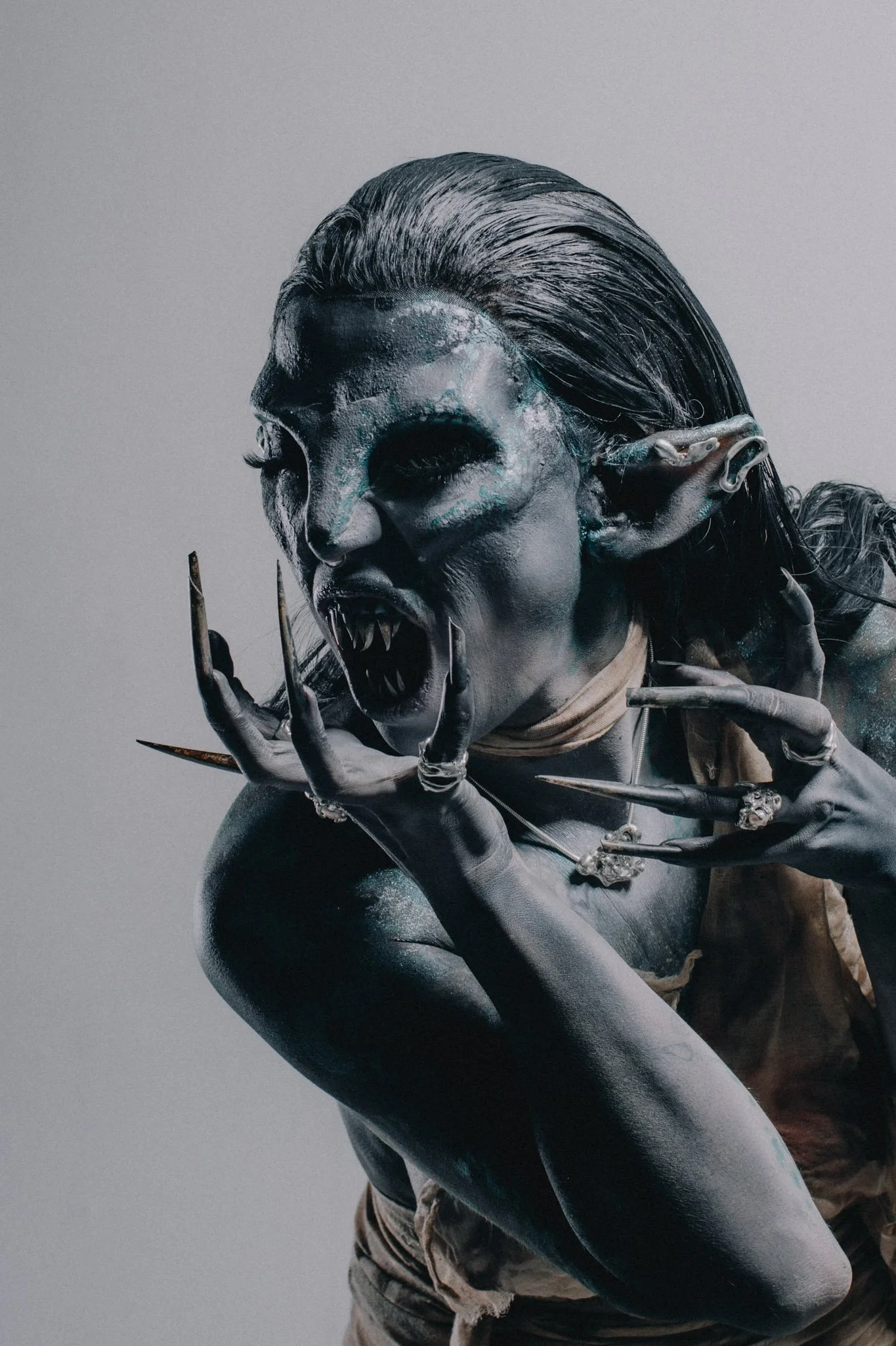 A person with face paint resembling a skull, wearing elf ear prosthetics, with long slicked-back hair, fierce expression, and sharp claws, against a plain background.