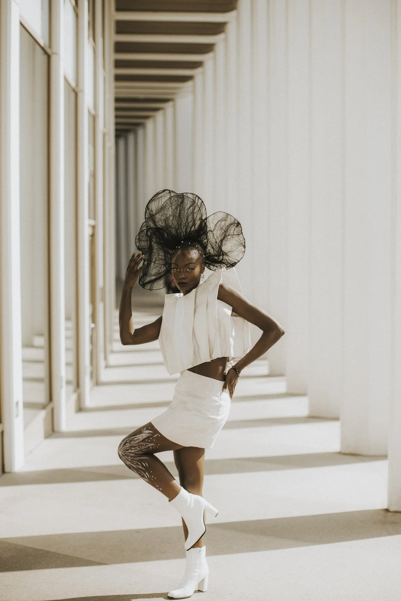 A fashion model with large black mesh hair and dark lipstick posing in a white outfit with bronze patterned tights and white boots in a modern, minimalist architectural setting.