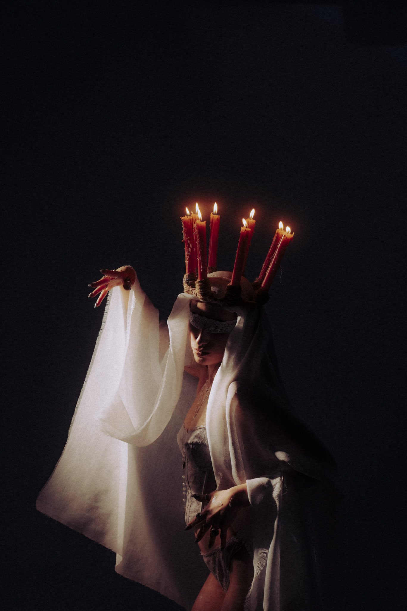 A person dressed as a deity or mythical figure with a white flowing garment and dramatic makeup, holding a staff topped with lit candles, against a dark background.