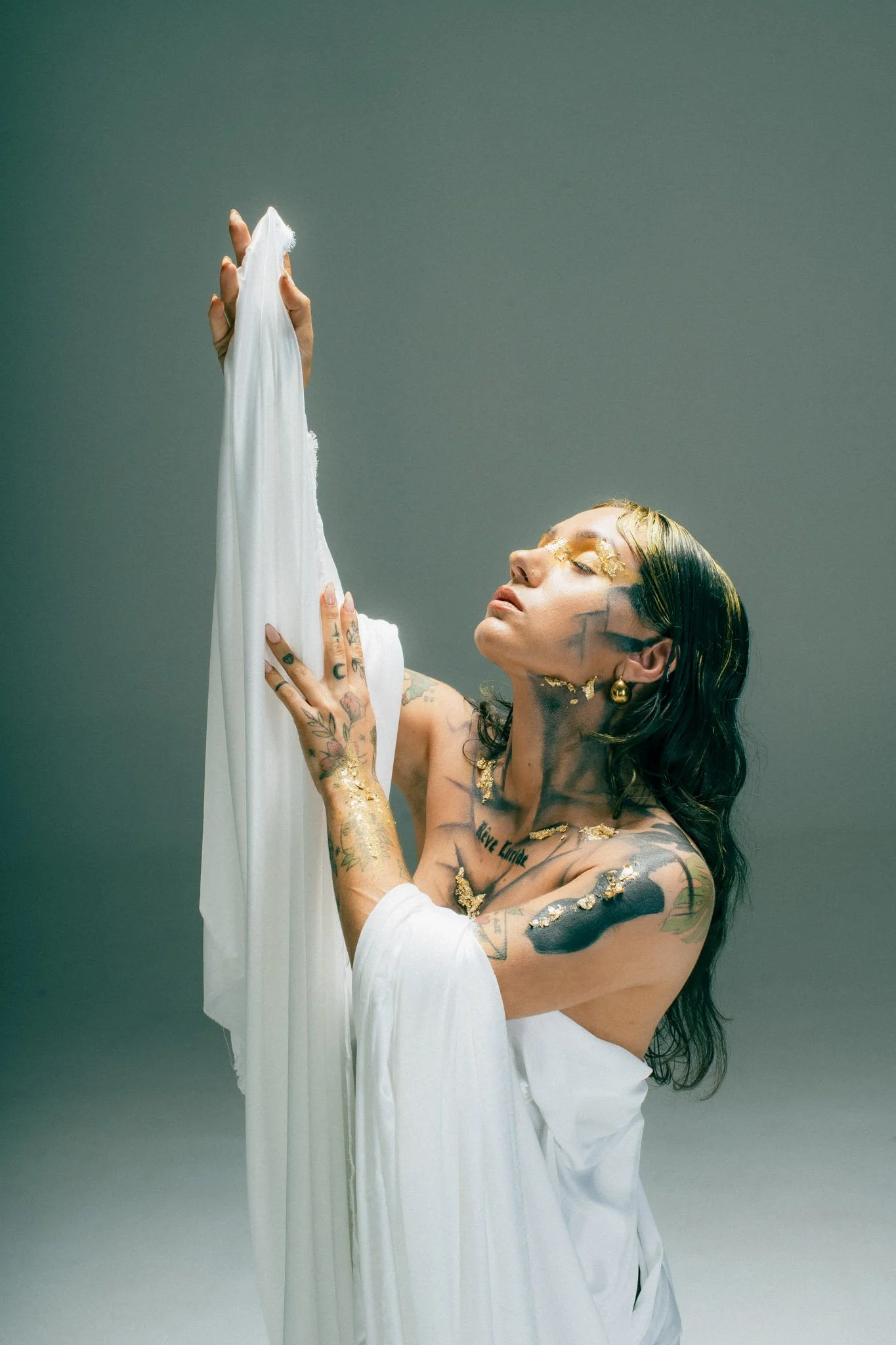 A woman with tattoos and gold leaf accents on her face and arms, wearing a white off-the-shoulder robe, holding a white fabric against a plain background.