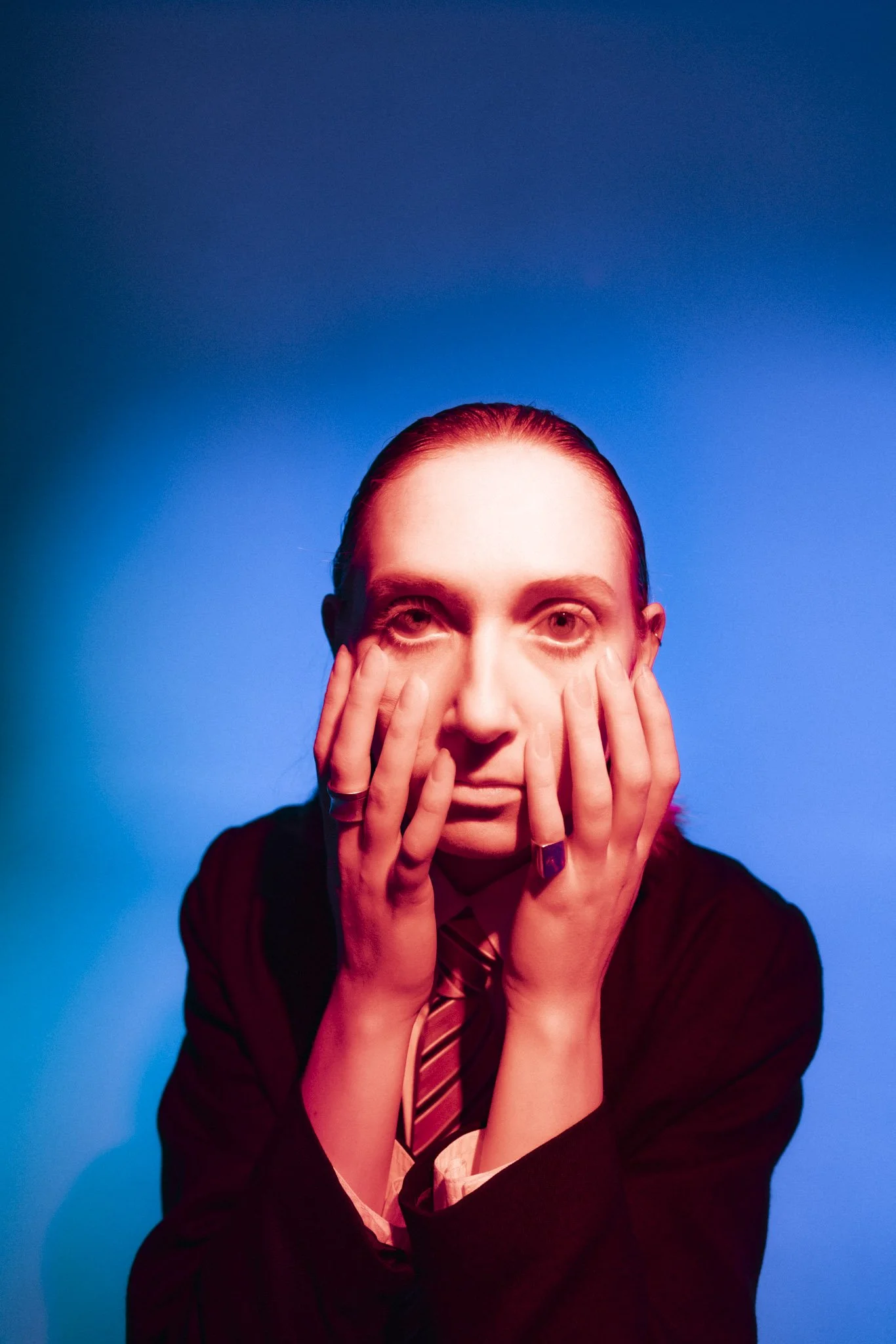 Person with red hair and light skin rubbing their face with both hands, wearing a dark blazer, white shirt, and a necktie, against a blue background.