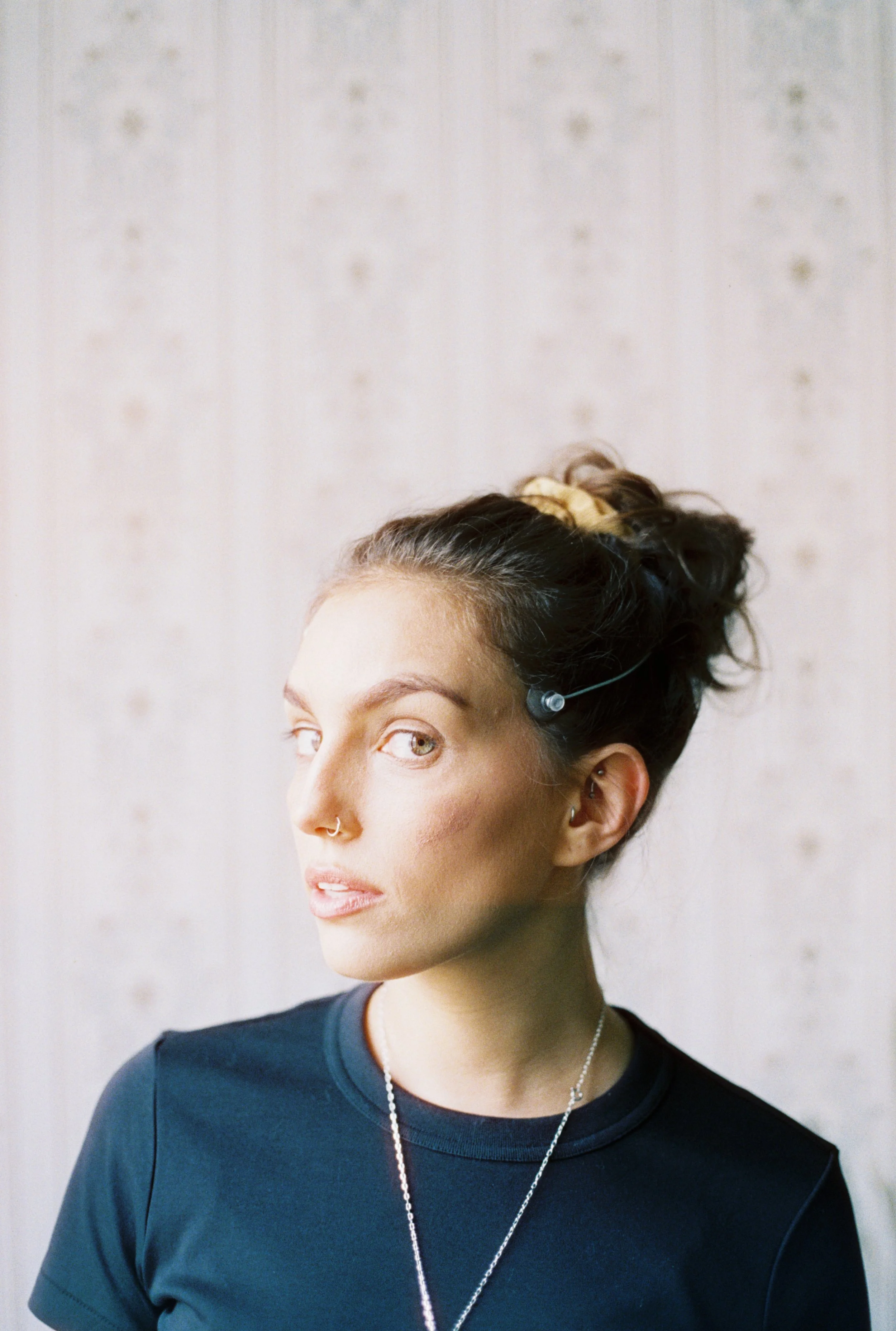 Close-up portrait of a woman with dark hair in a bun, wearing a blue t-shirt, silver necklace, and earrings, looking to the side against a light background.