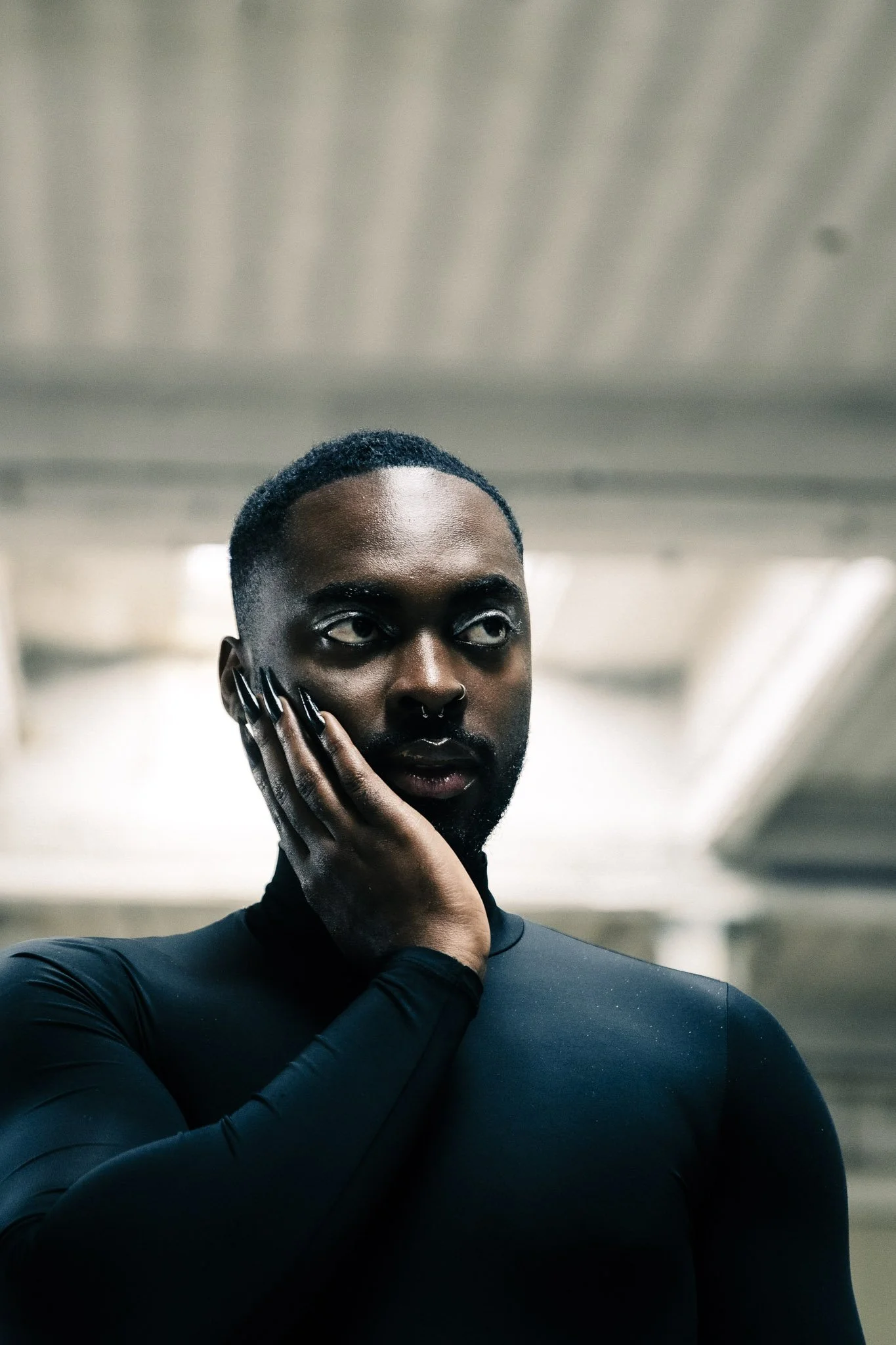 A close-up of a person with short black hair and dark skin, touching their face with a hand, wearing a black long-sleeve top, in an indoor setting with blurred background.