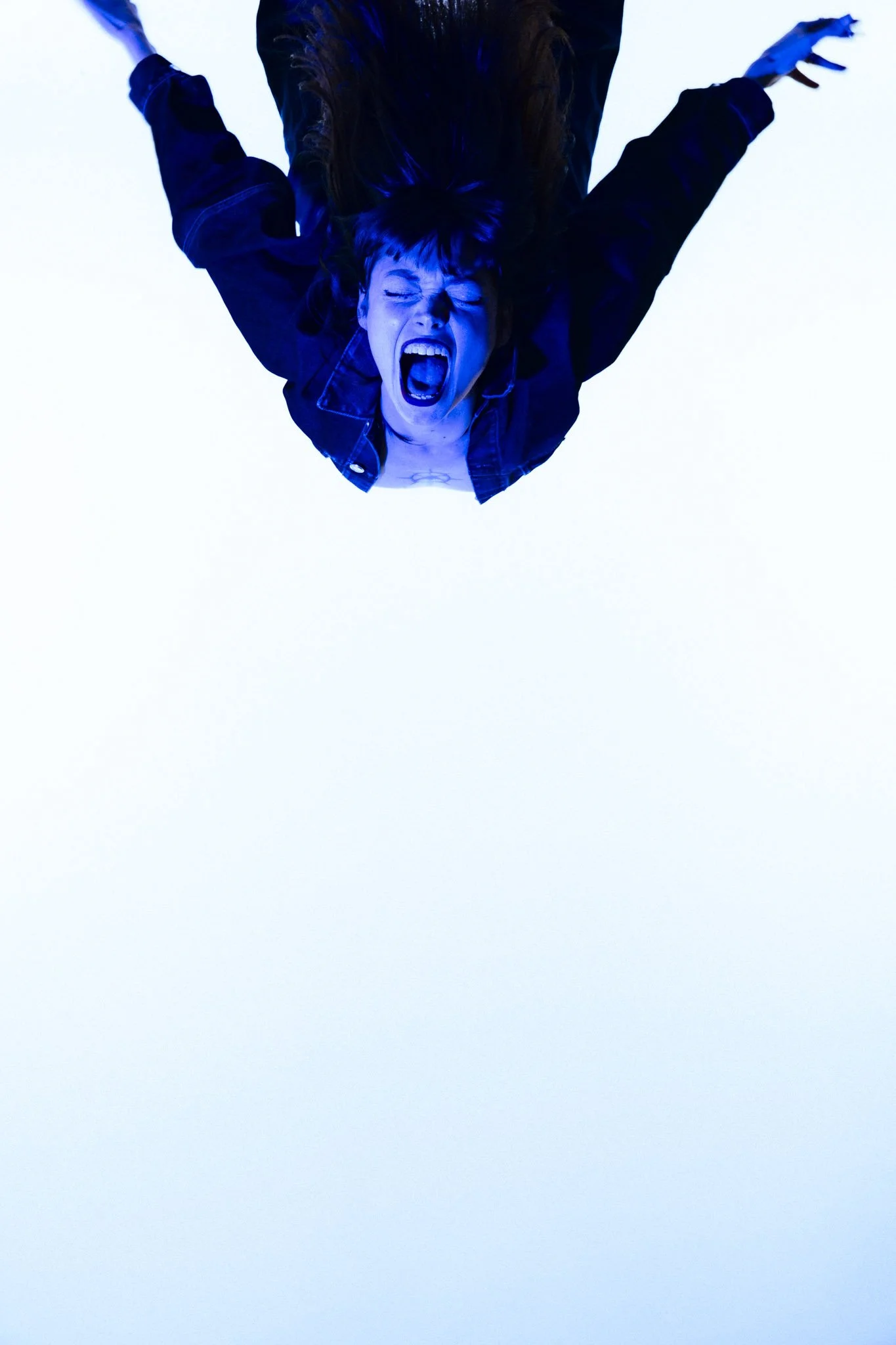 A woman with dark hair and wearing a black jacket is captured from above, with her mouth open as if shouting or singing. She is lying on a white surface, illuminated with blue light.