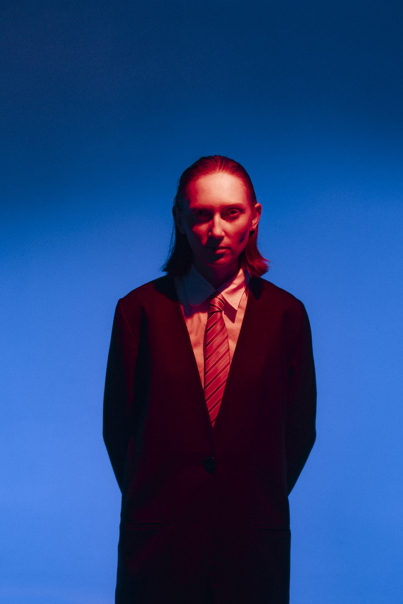 A woman with red hair dressed in a black blazer, white shirt, and striped red tie stands against a blue background, illuminated by red and blue lighting.