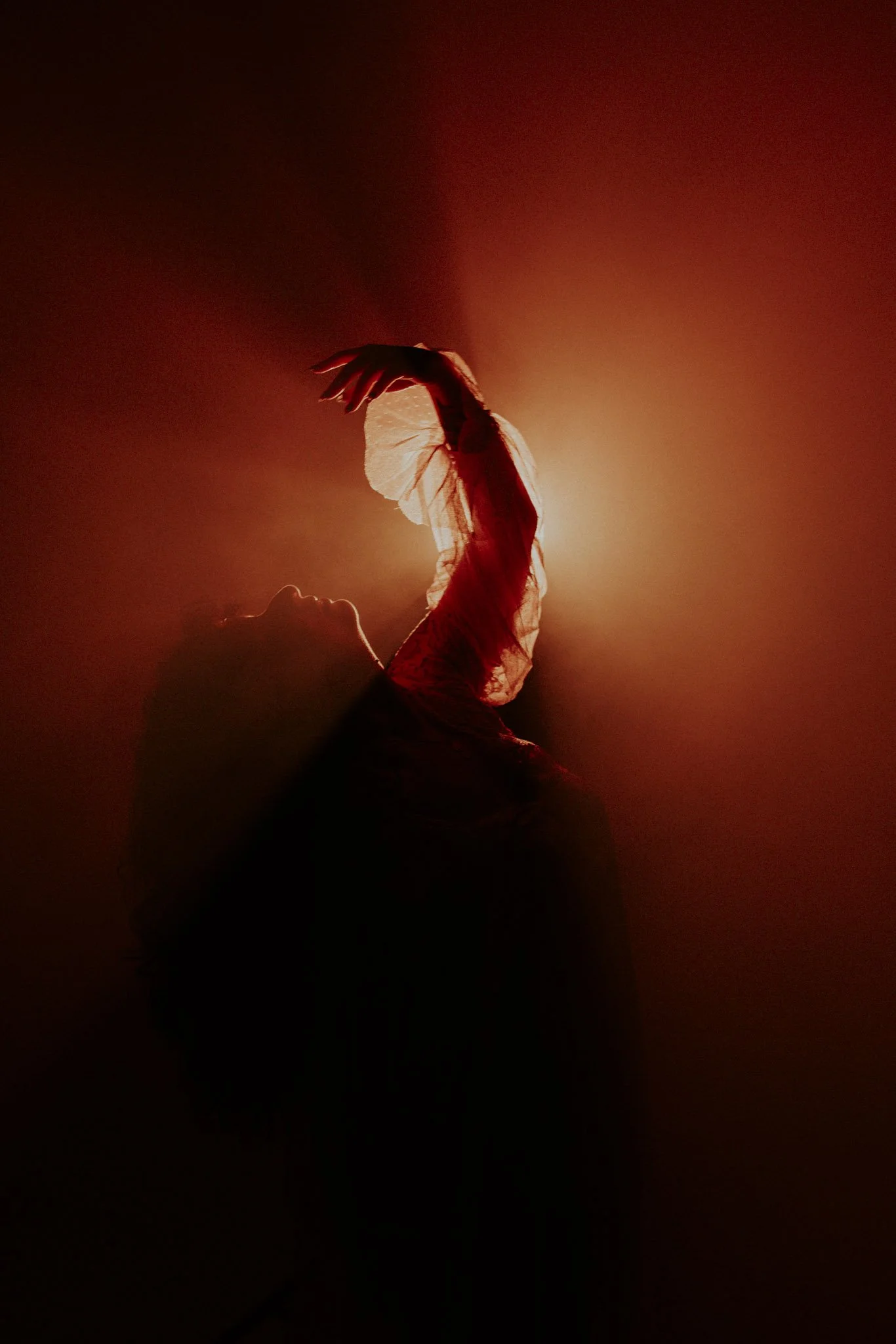 A person is lying on their back with their arm raised, illuminated by a warm, orange-red light, creating a silhouette against a dark background. The person appears to be wearing a translucent, flowing fabric.