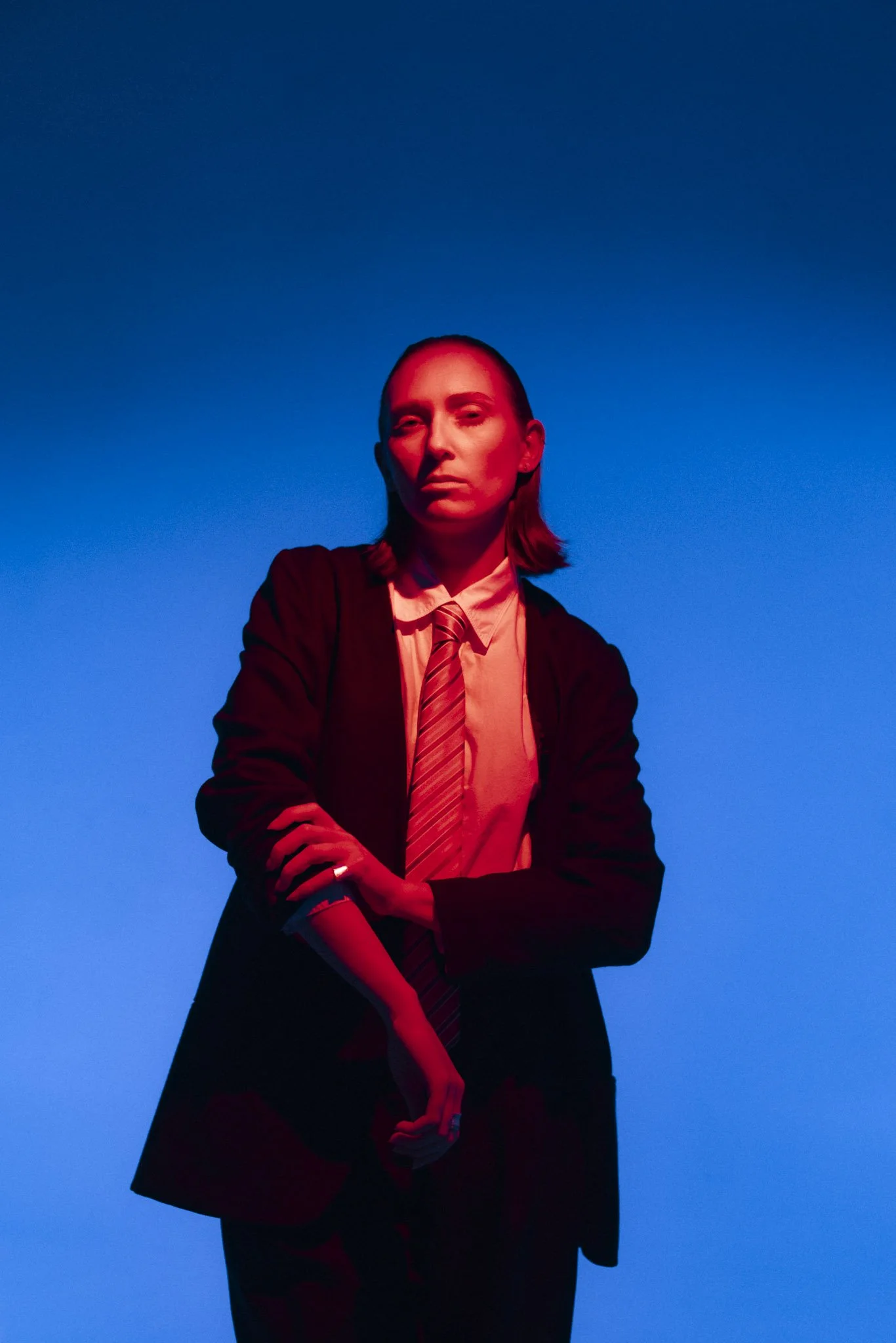 A young woman with long hair, dressed in a suit, white shirt, and striped tie, stands against a gradient blue background illuminated with red lighting. She has a confident expression with her eyes slightly closed and her arms crossed.