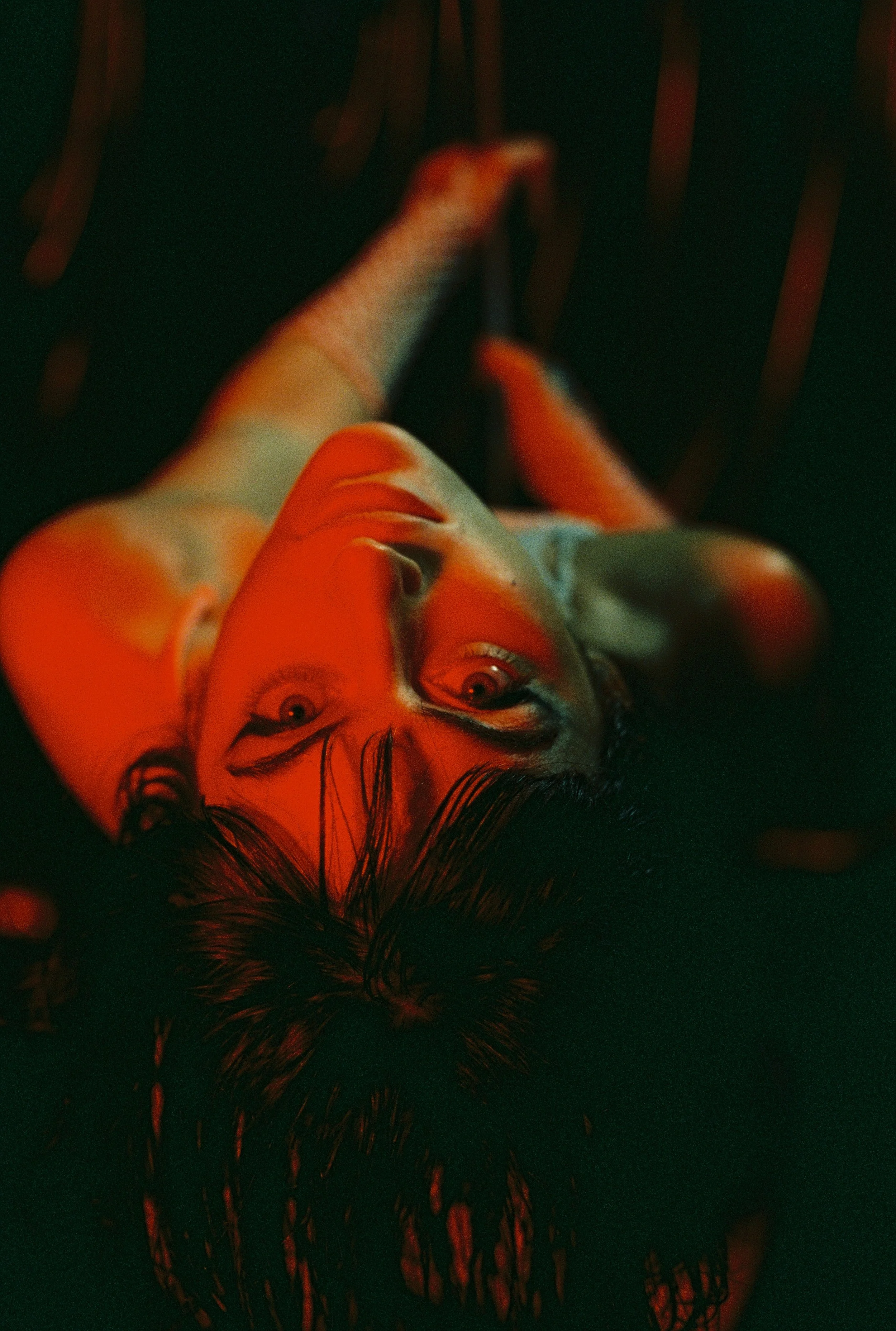 A woman with dark hair and tattoos on her arm, lying on her back, looking up, with red lighting highlighting her face and tattoos, against a dark background.