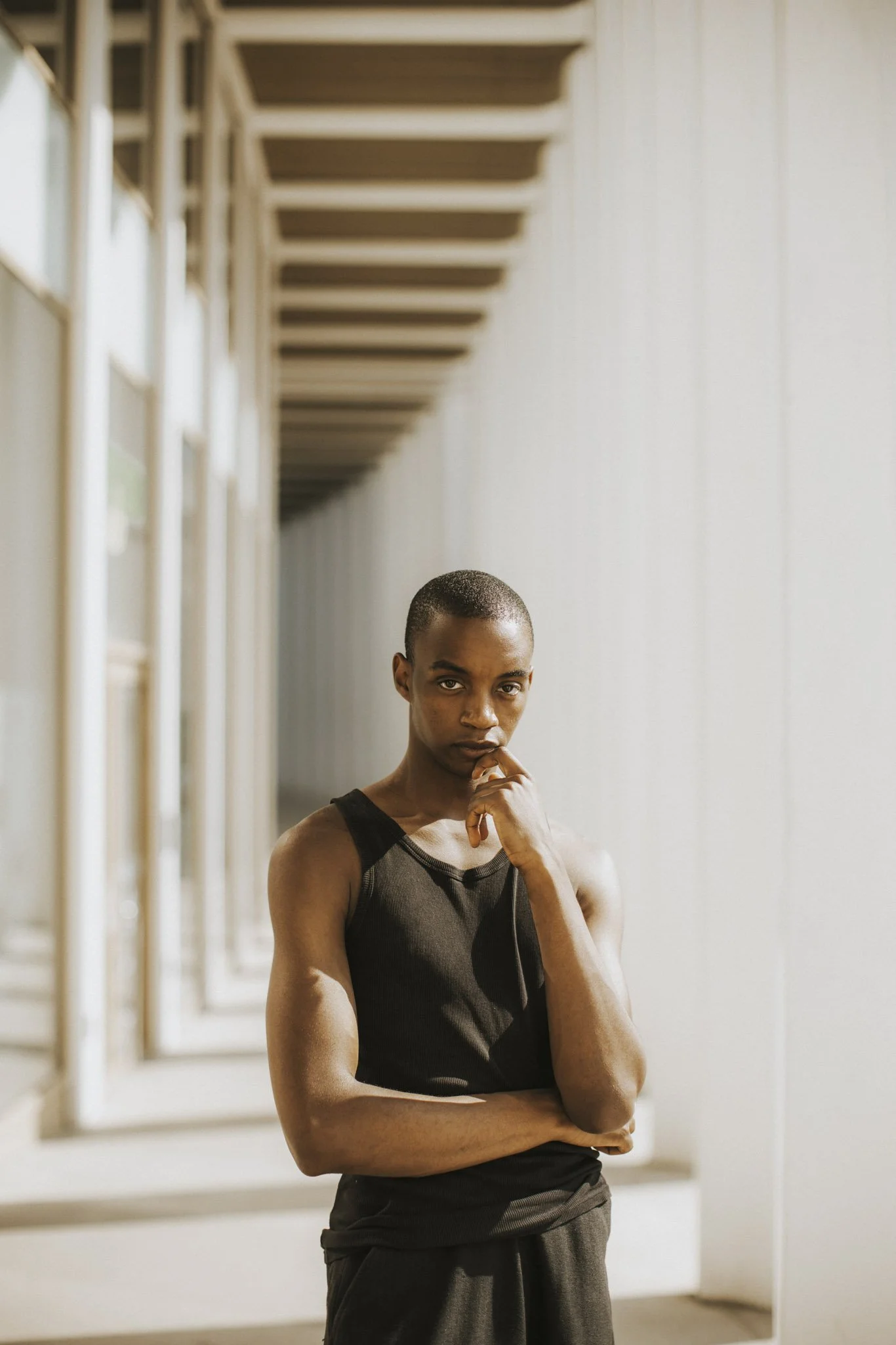 A young man with a shaved head wearing a black sleeveless athletic shirt and dark pants standing indoors against a white wall with a row of windows. He has one hand resting on his chin and is looking directly at the camera.