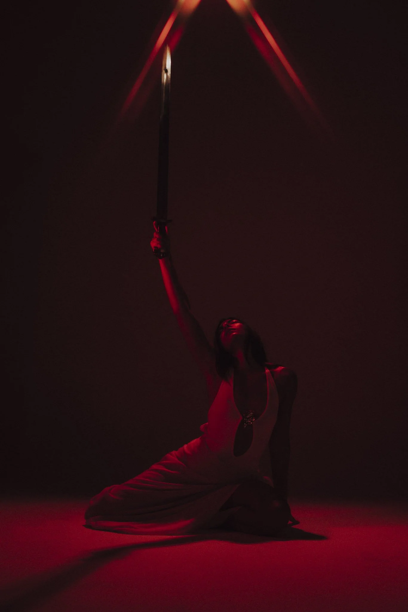 A woman dressed in white dancewear performs a martial arts pose with a sword, illuminated by red lighting in a dark setting.