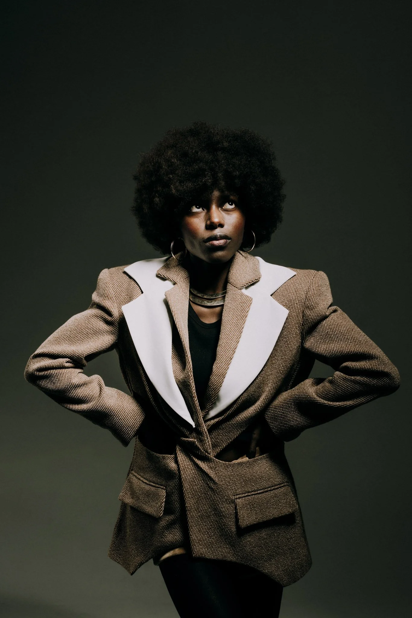 A woman with dark skin and a large afro hairstyle wearing a brown and white checkered blazer and black top, standing against a dark background.
