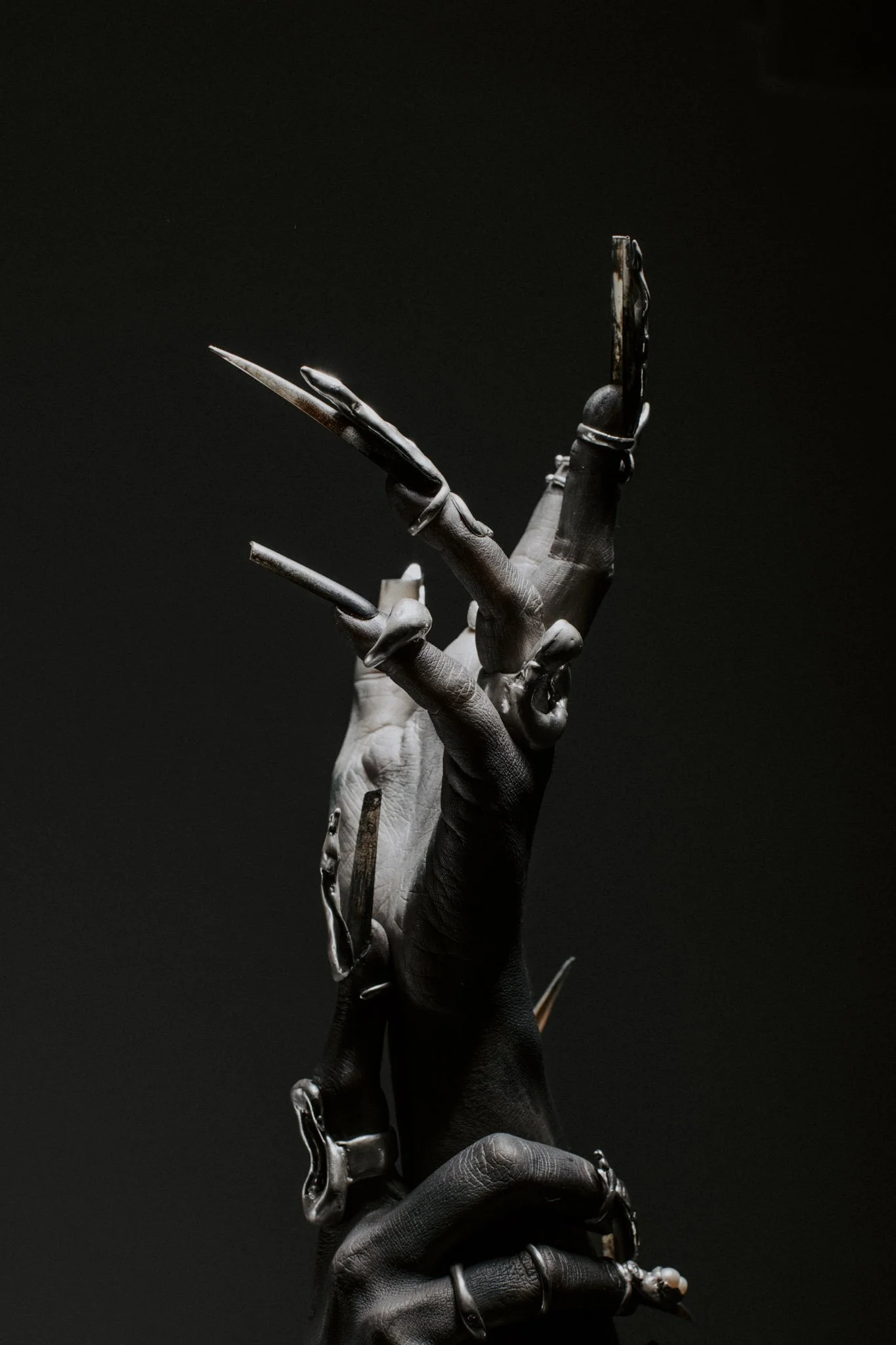 A black and white photograph of a skeletal hand with long, pointed nails, holding a rusted metal object against a dark background.