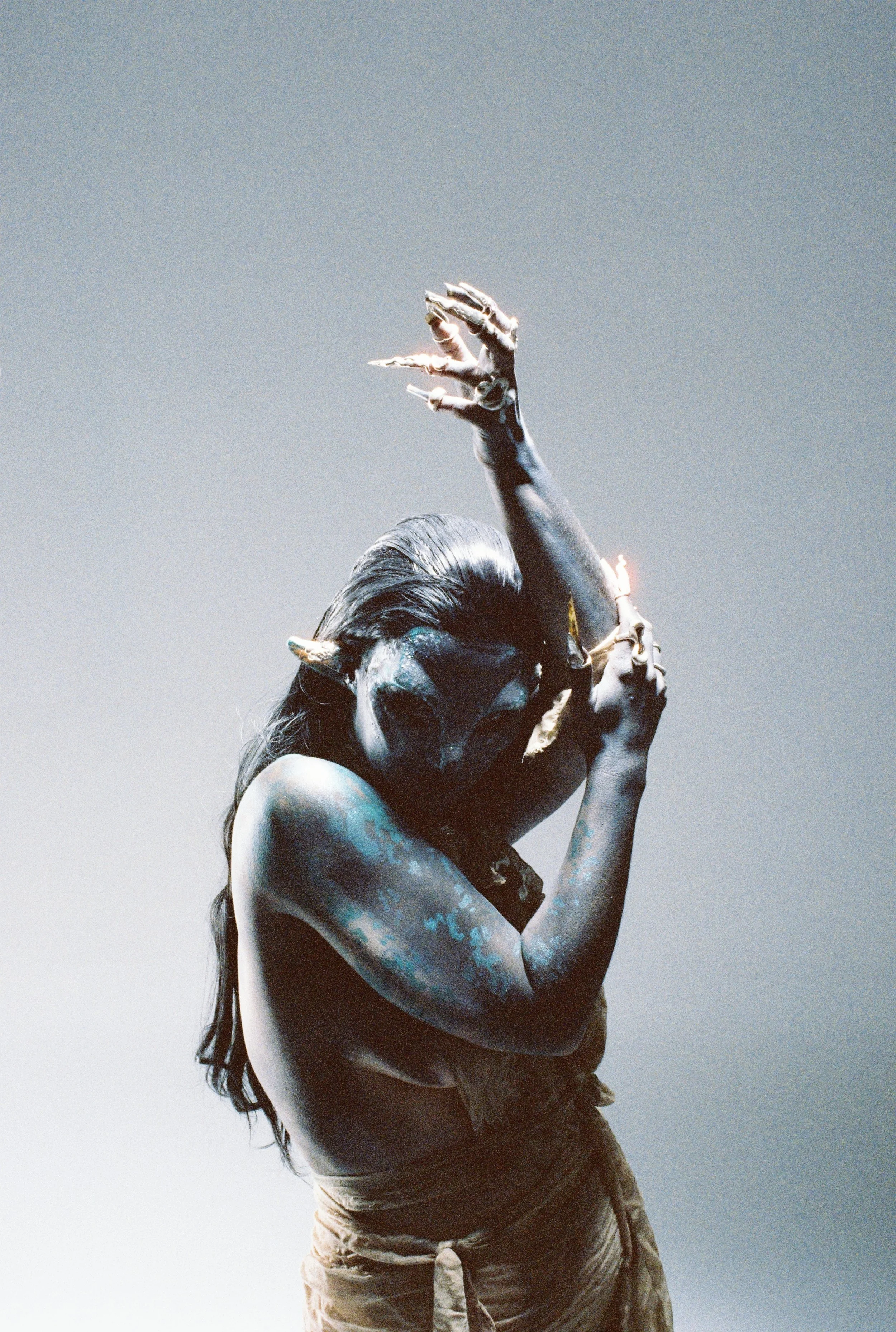 A person with painted skin and long hair, posing with arms raised and head tilted, against a plain background.