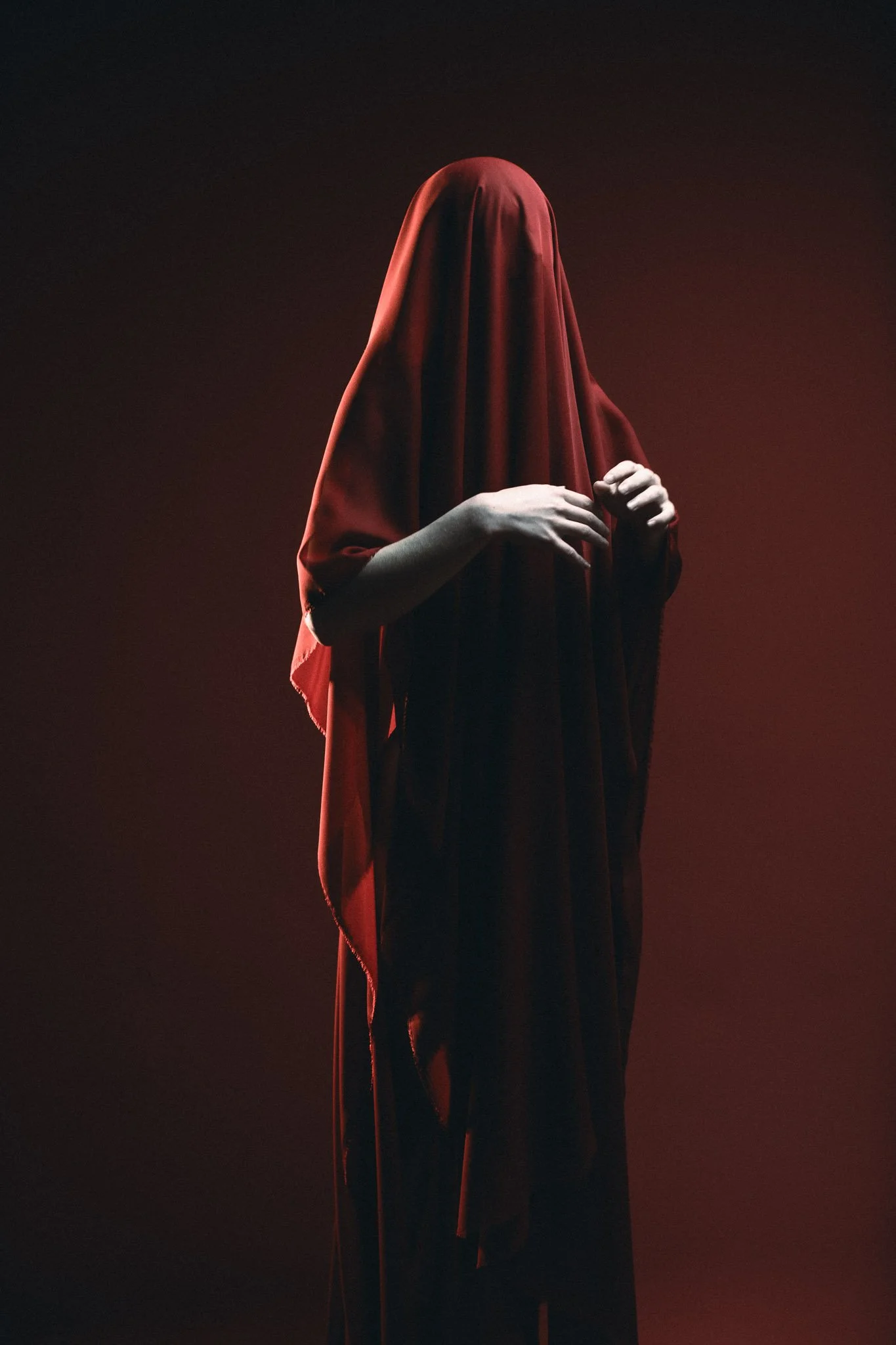 Person standing with a red veil covering their head and face, holding a thin cane in their right hand, against a dark gradient background.