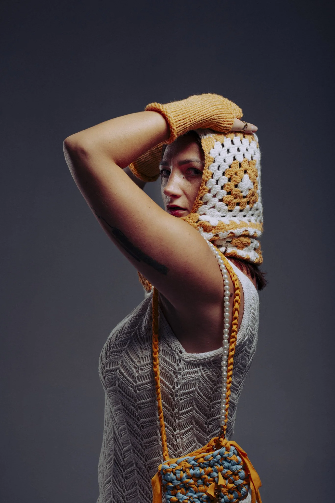 A woman with light skin and dark hair wearing a cream-colored lace top and a knitted yellow and white scarf, accessorized with a pearl necklace, orange knitted gloves, and a multicolored knitted purse, poses with her arm raised and hand resting on he