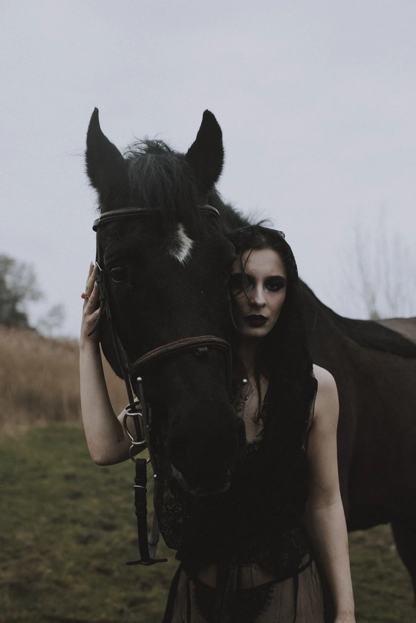 A woman dressed in black gothic attire holding a black horse in an outdoor setting during overcast weather.