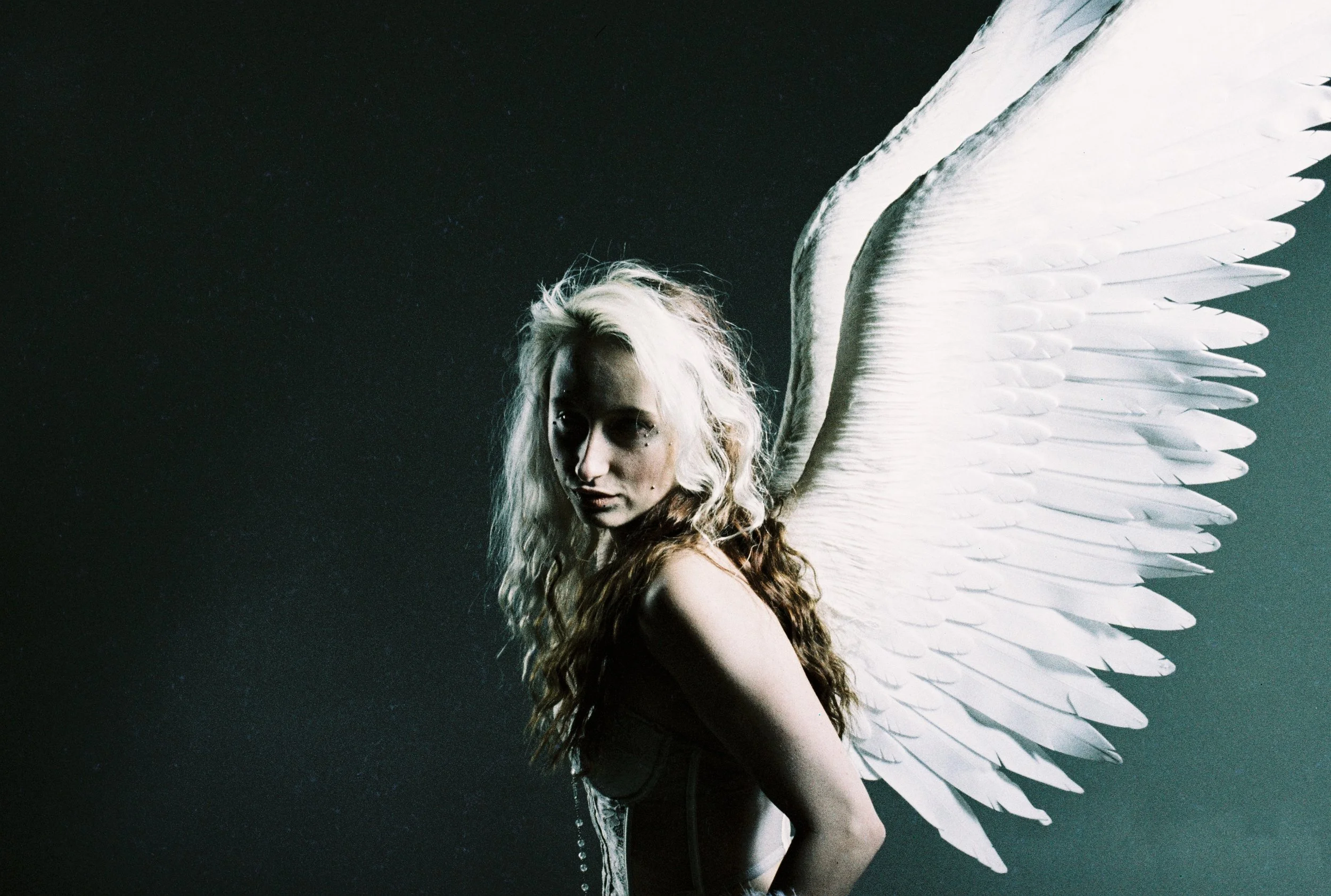 A woman with long, wavy blonde hair, wearing a black dress with lace, is standing with her arms crossed, against a dark background. She has large white wings attached to her back.