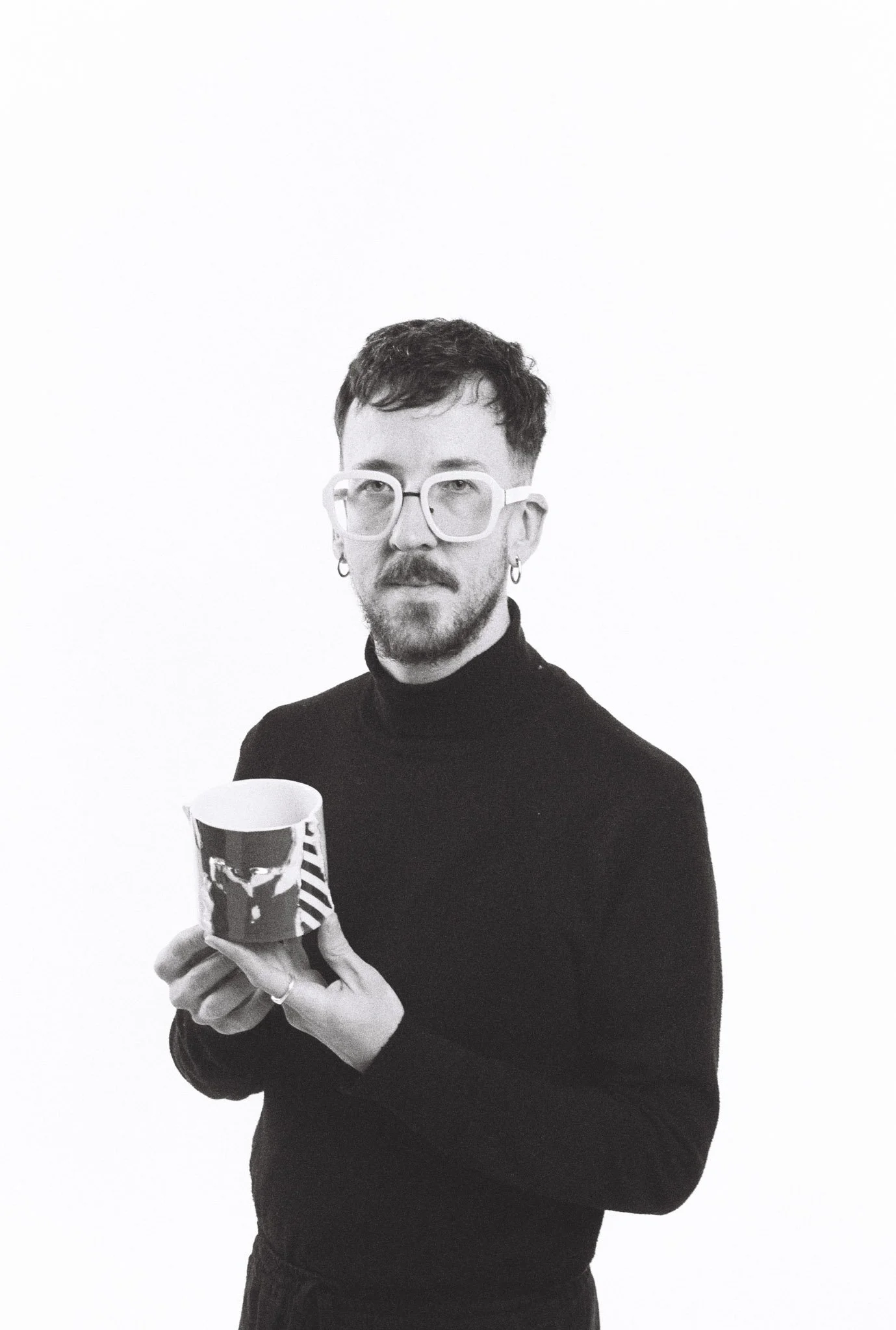 Black and white photograph of a man with short dark hair, glasses, and earrings, wearing a black turtleneck sweater, holding a striped mug with a bold design.