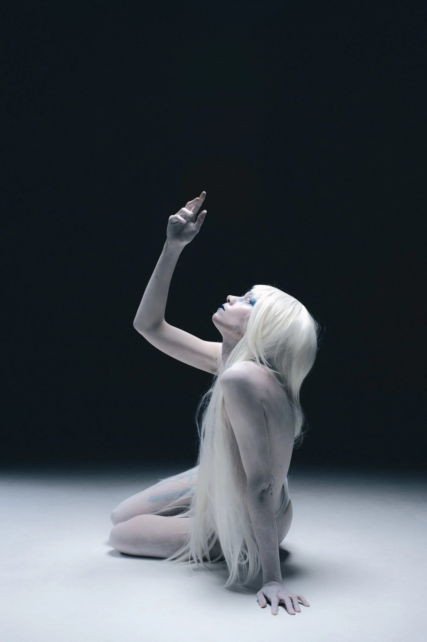 A person with long platinum blonde hair kneels on the ground, supporting themselves with one hand, with a dark background.