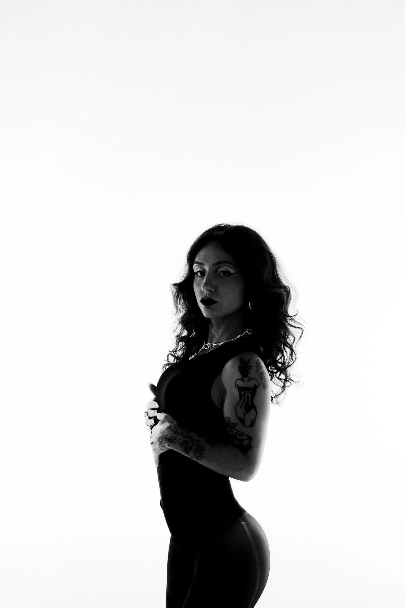 Black and white photo of a woman with tattoos on her arms, looking at the camera, with a high ponytail, wearing a sleeveless top and fitted pants.