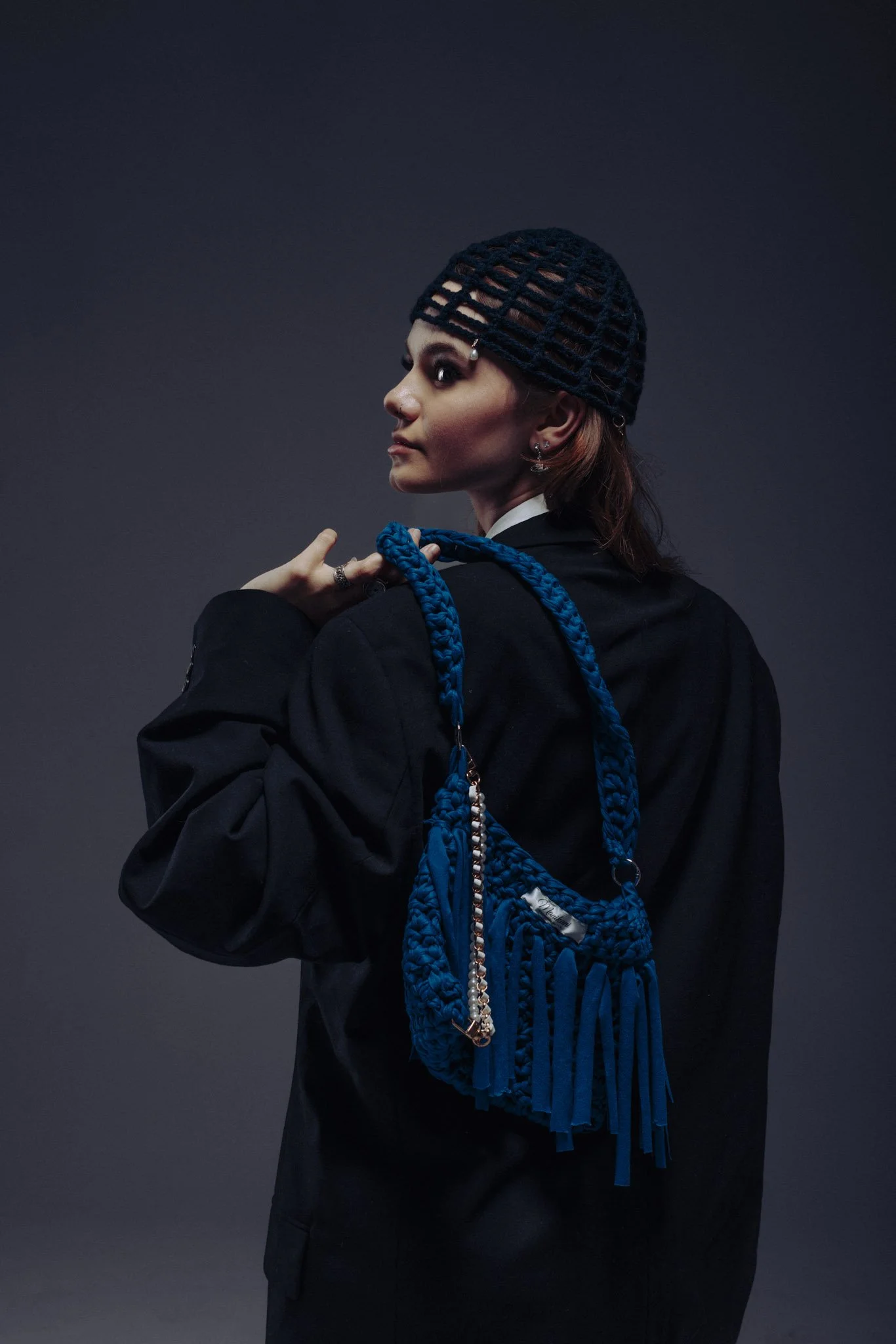 A woman with red hair wearing a black wool cap with a grid pattern, a black blazer, and accessorized with earrings, holding a blue woven bag with a chain strap against a dark background.
